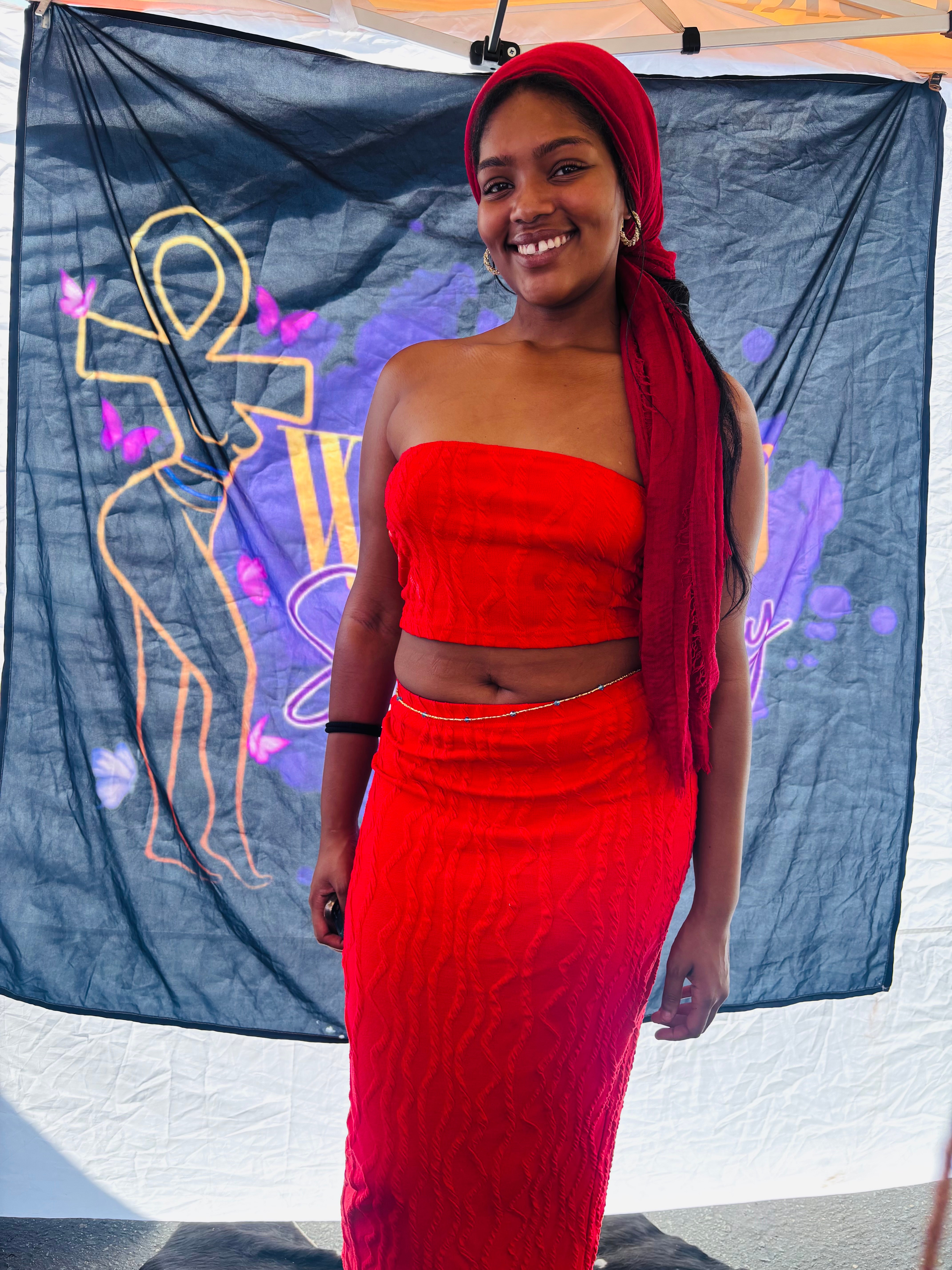 Young woman in red strapless top and skirt smiling, standing in front of a colorful backdrop with abstract designs.