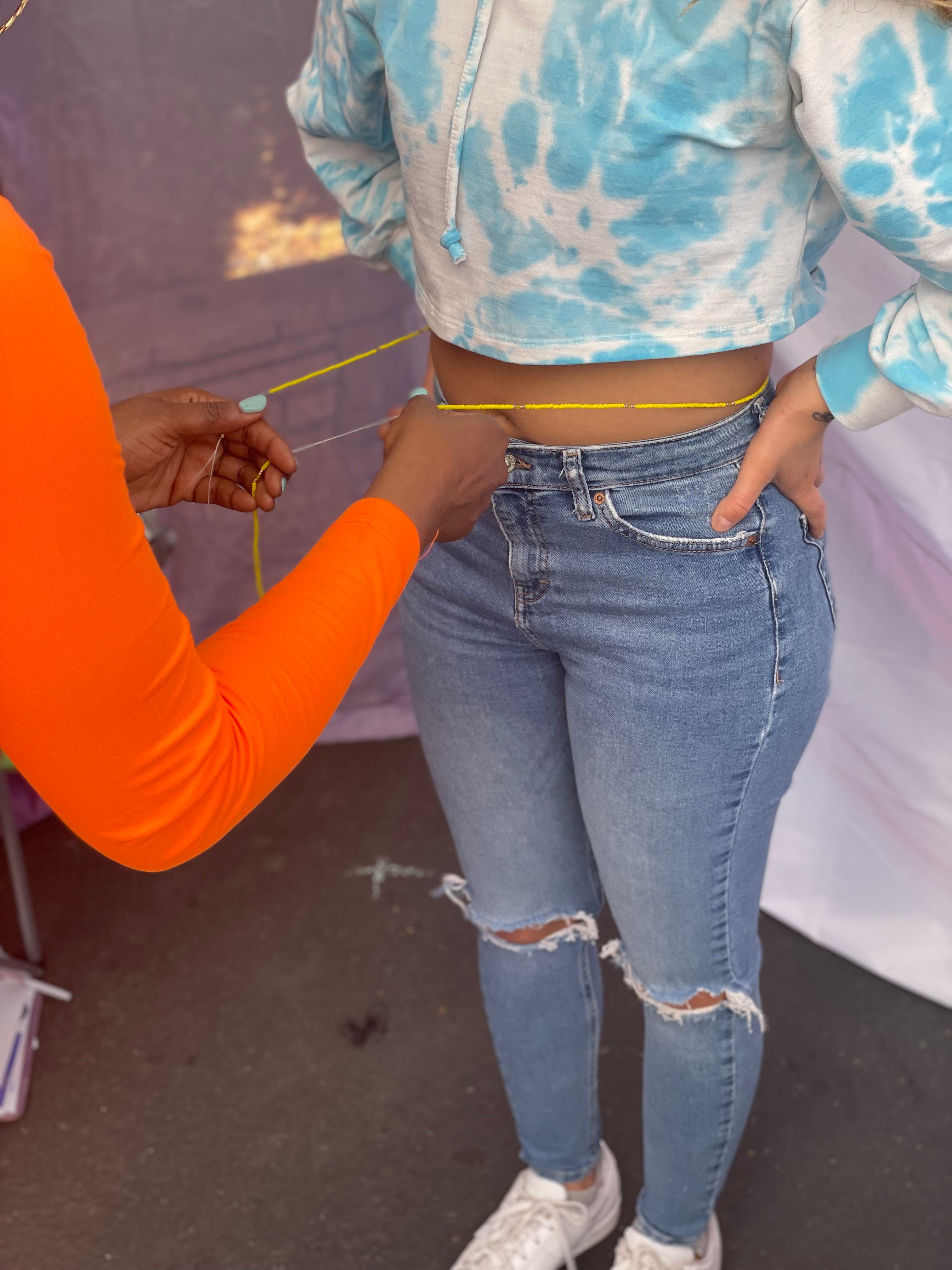 Person measuring waist of another person wearing a blue tie-dye top and ripped jeans, outdoors.