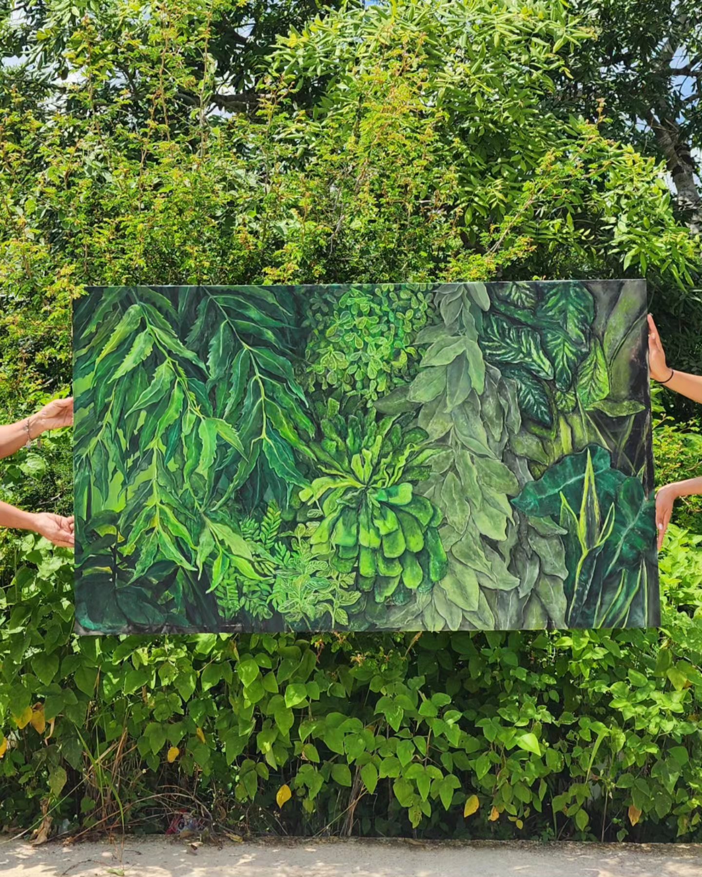 Two people hold a painting of green leaves and plants outdoors, with trees and bushes in the background.