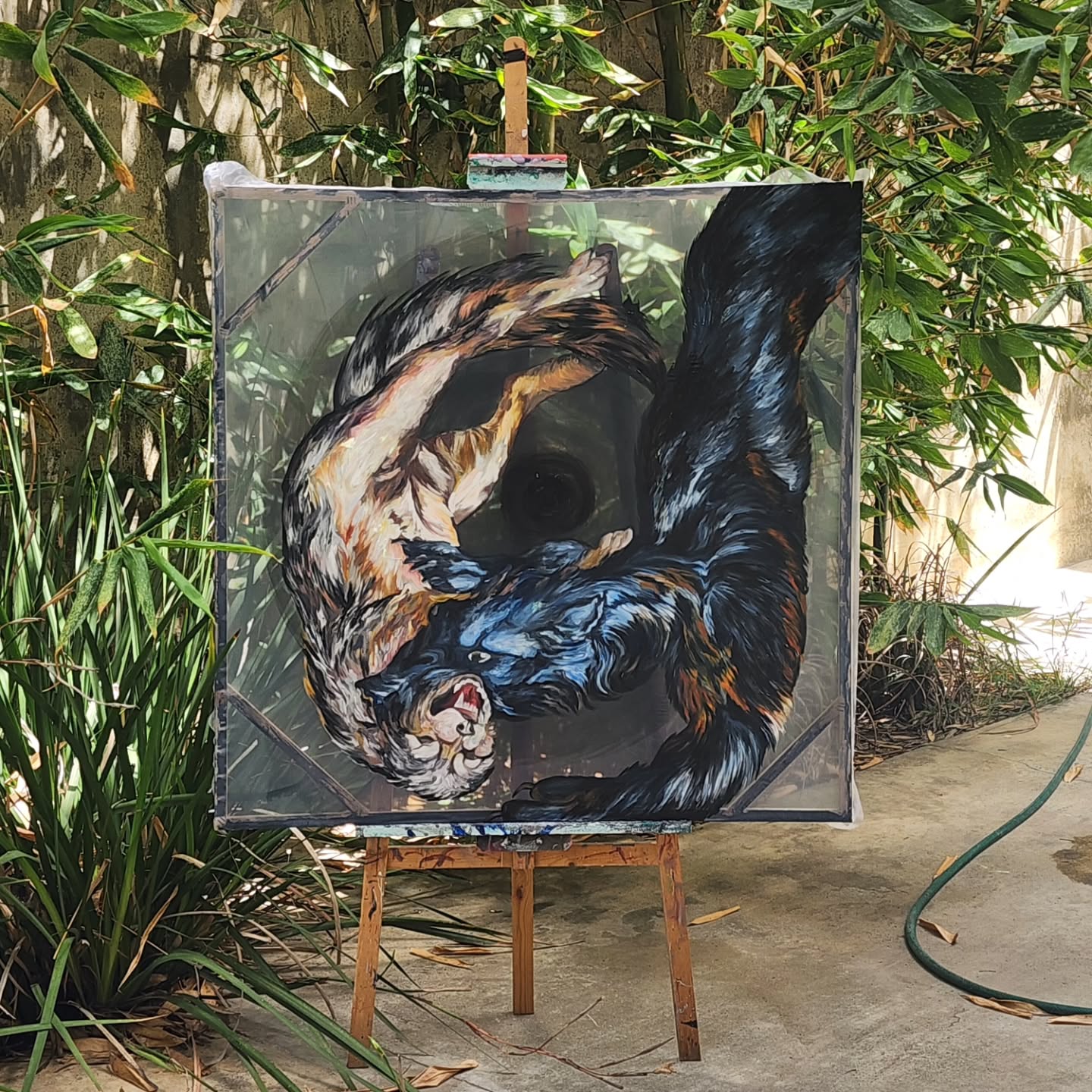 Painting of a tiger lying on its side, with detailed fur and facial features, on an easel outdoors surrounded by greenery.