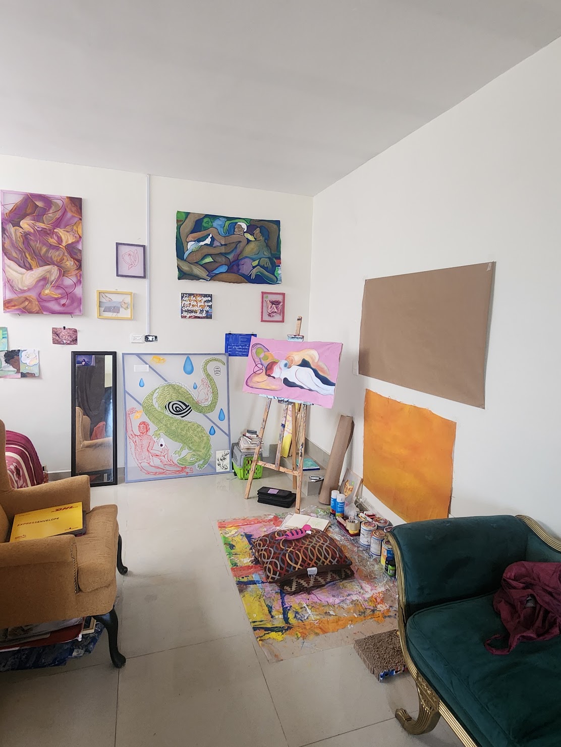 Art studio with paintings, a mirror, and furniture including a yellow armchair and green sofa, on a colorful rug.