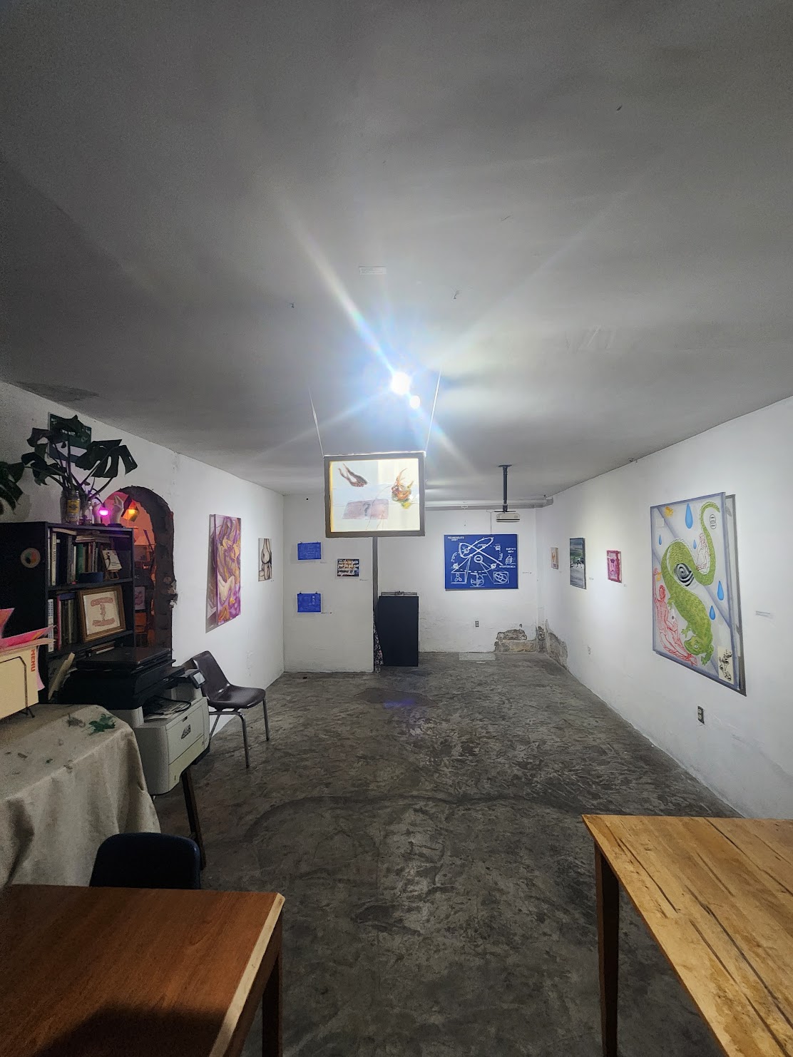 Gallery room with paintings on white walls, concrete floor, and a bright light on ceiling. Furniture includes tables and chairs.