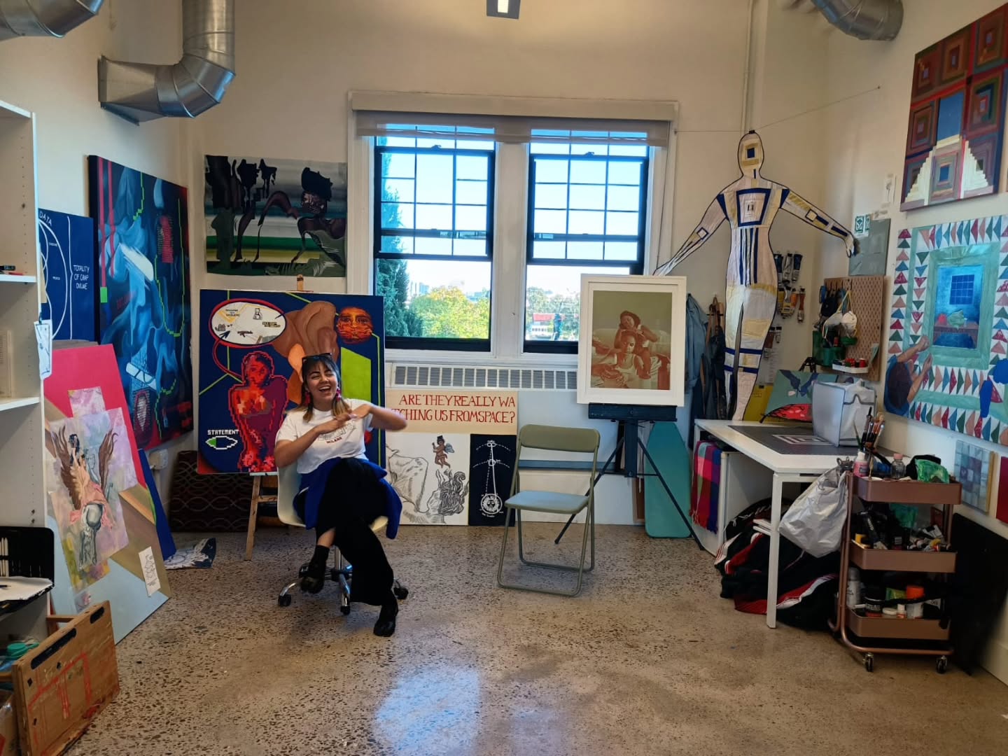 Person sitting on a chair in an art studio, surrounded by paintings, art supplies, and windows with a view outside.