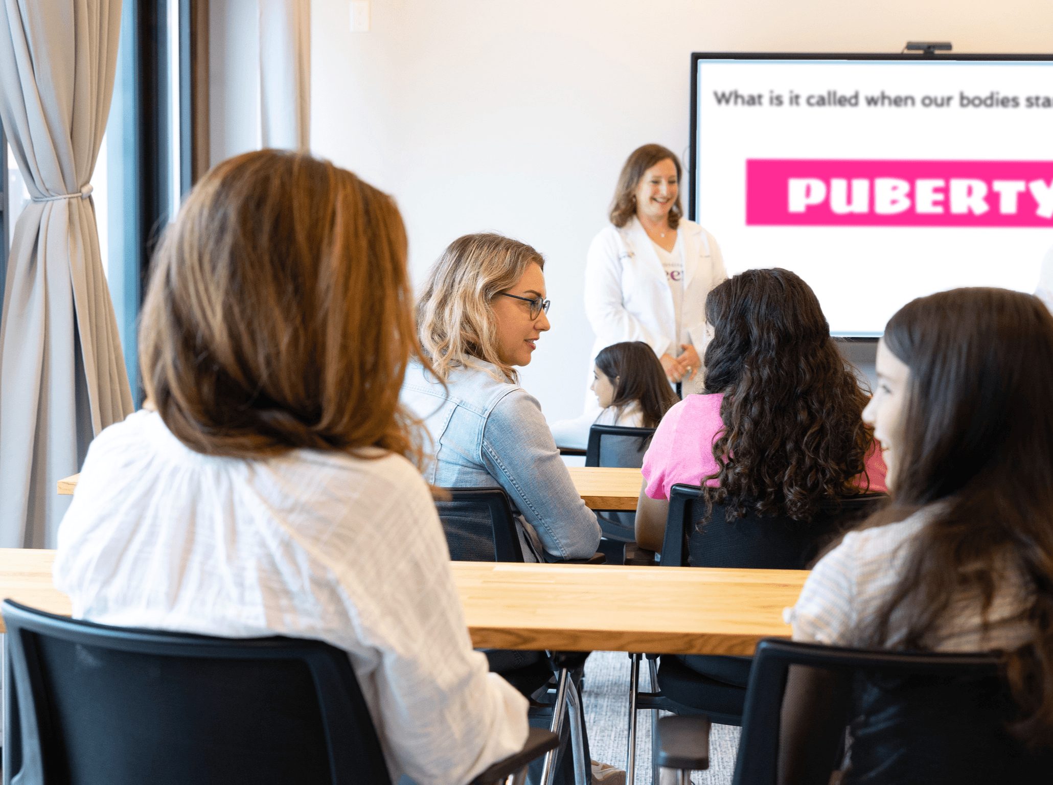 A classroom with a teacher and students, some looking at a presentation screen, in a well-lit room.