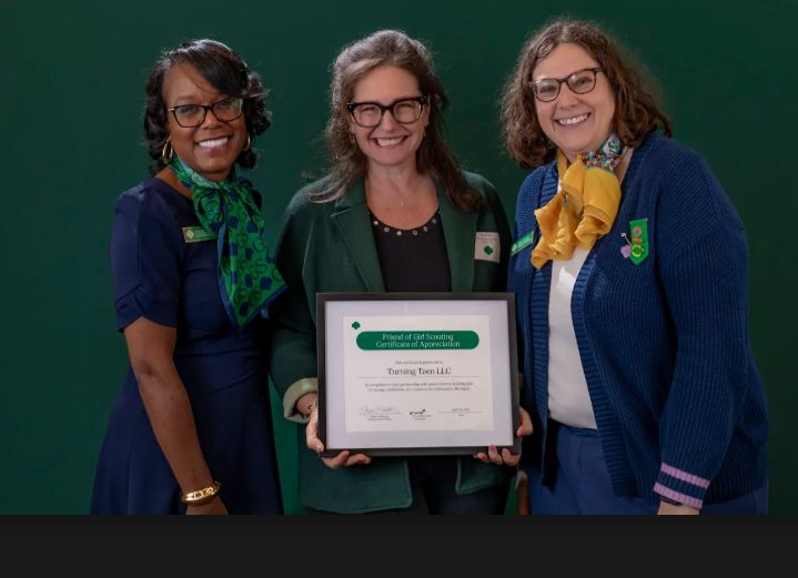 Three women standing together, one holding a framed certificate, smiling, against a green background.
