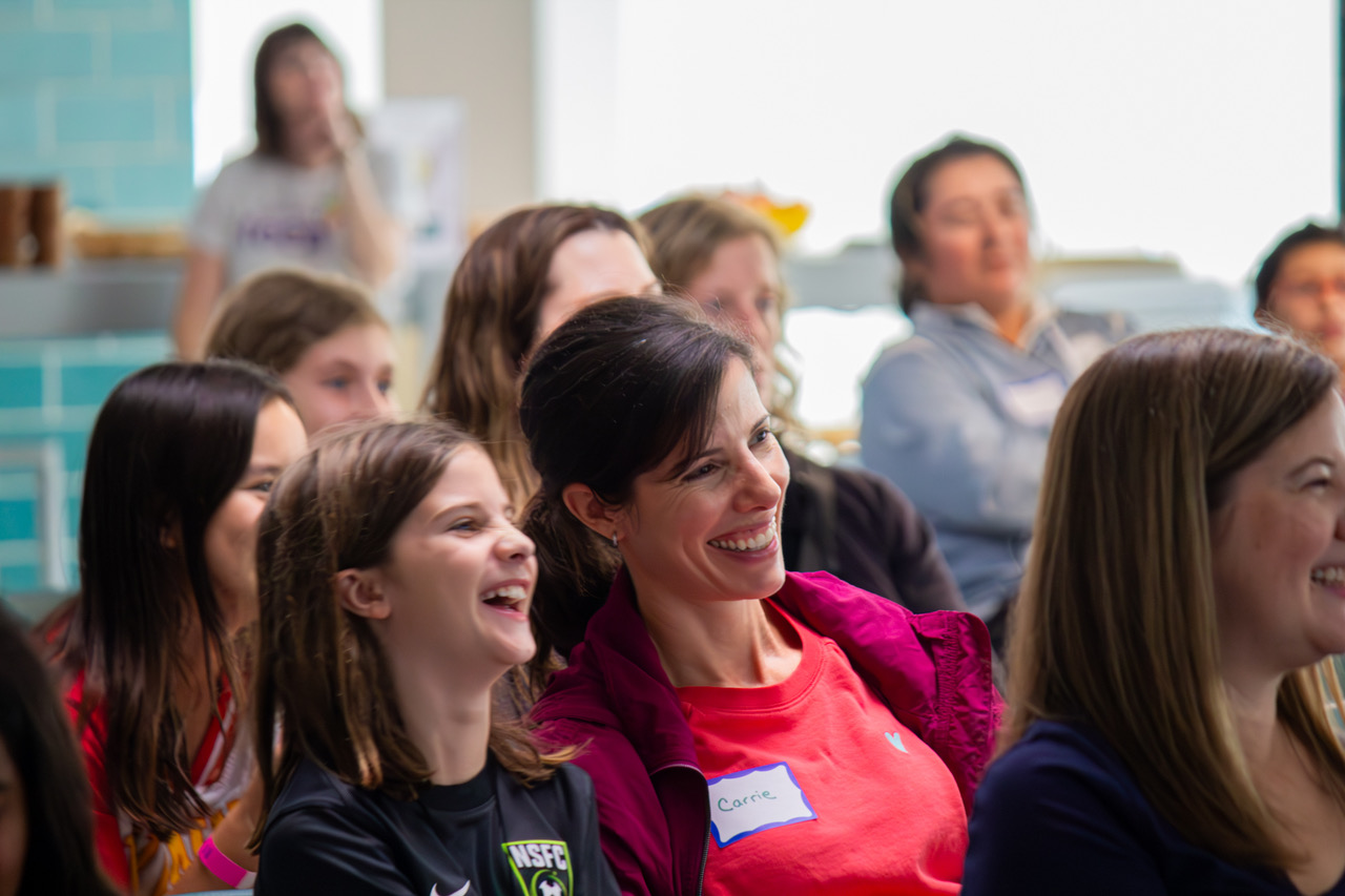 Group of smiling women and children sitting together indoors, engaging in a social event or gathering.