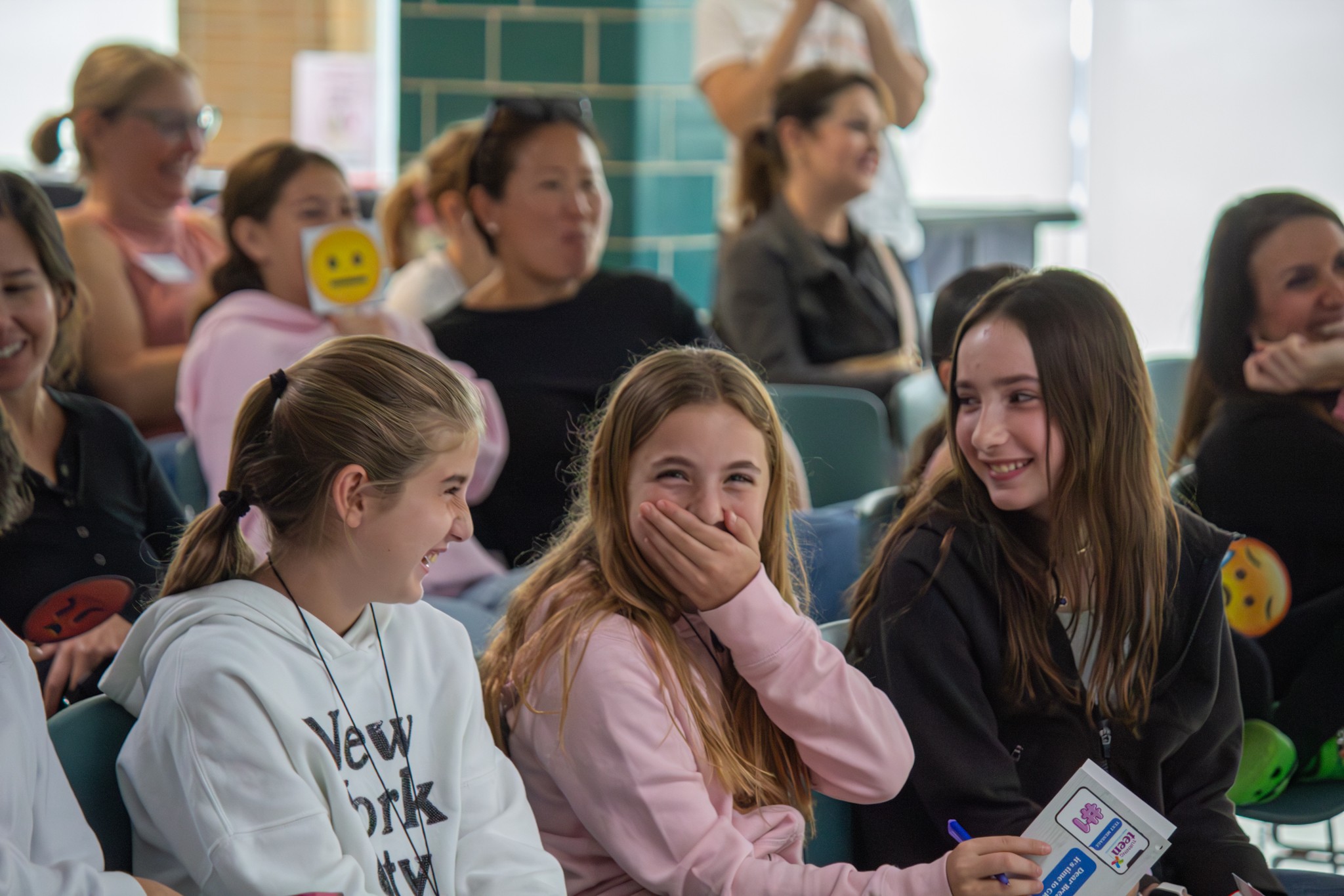Group of young women sitting and laughing in a bright room, some with emoji stickers on their clothing.
