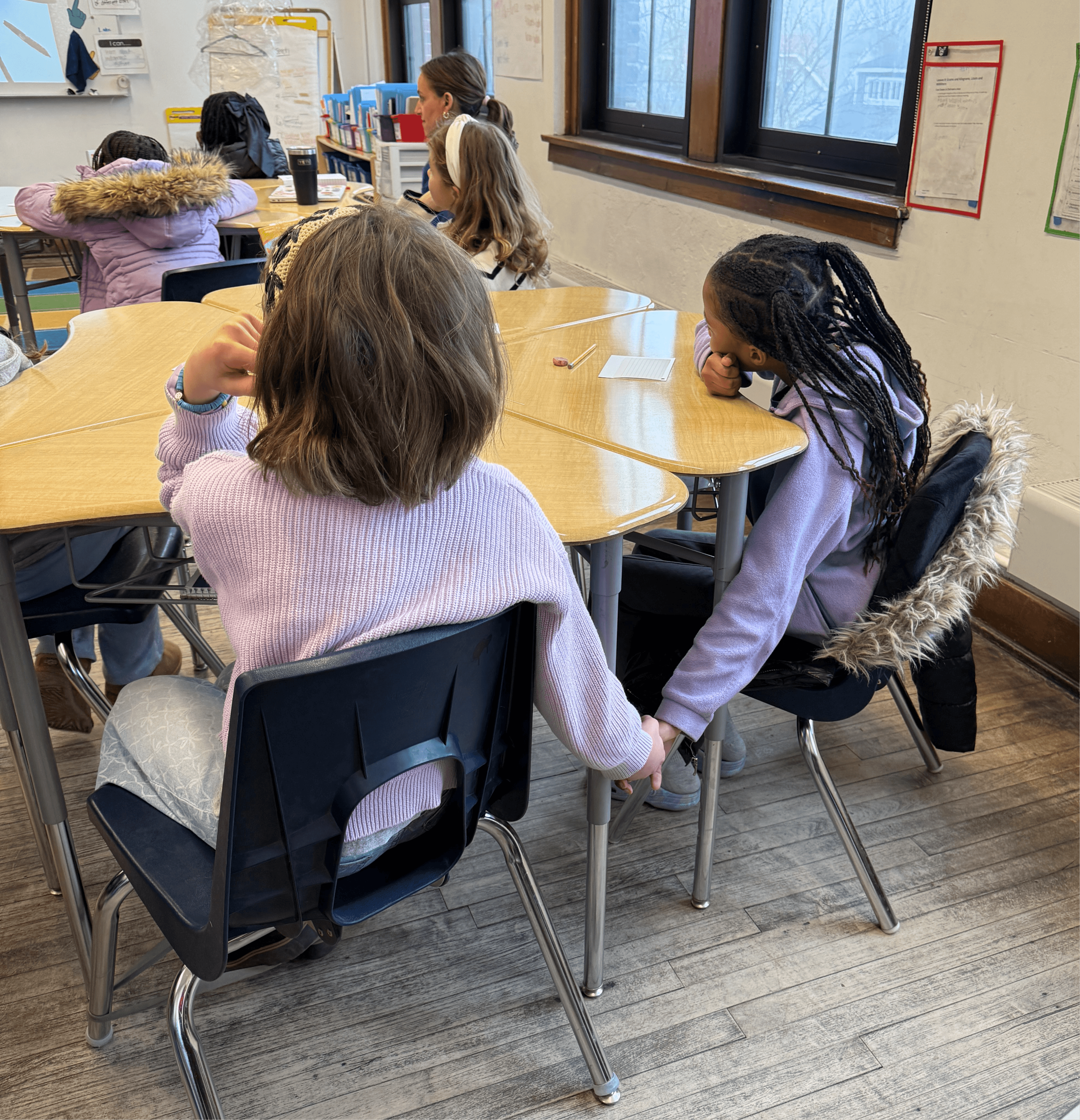 Two children sit at a table holding hands in a classroom, with other students and educational posters visible in the background.