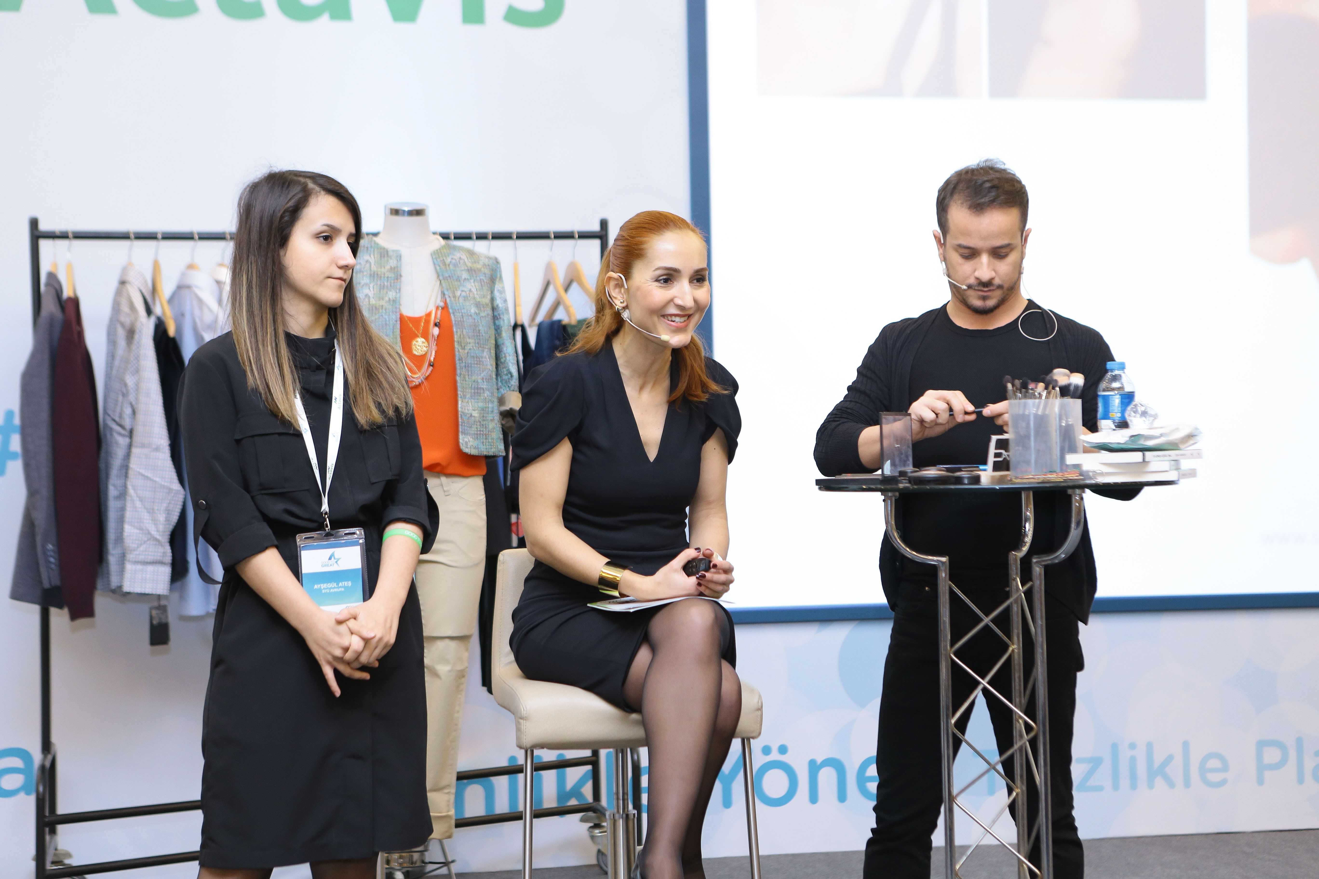 Three women at a presentation or event, one standing and two seated, with clothing racks in the background.