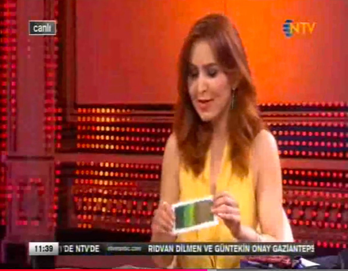 Woman with long hair in a yellow top holding a card on a TV set with red background.