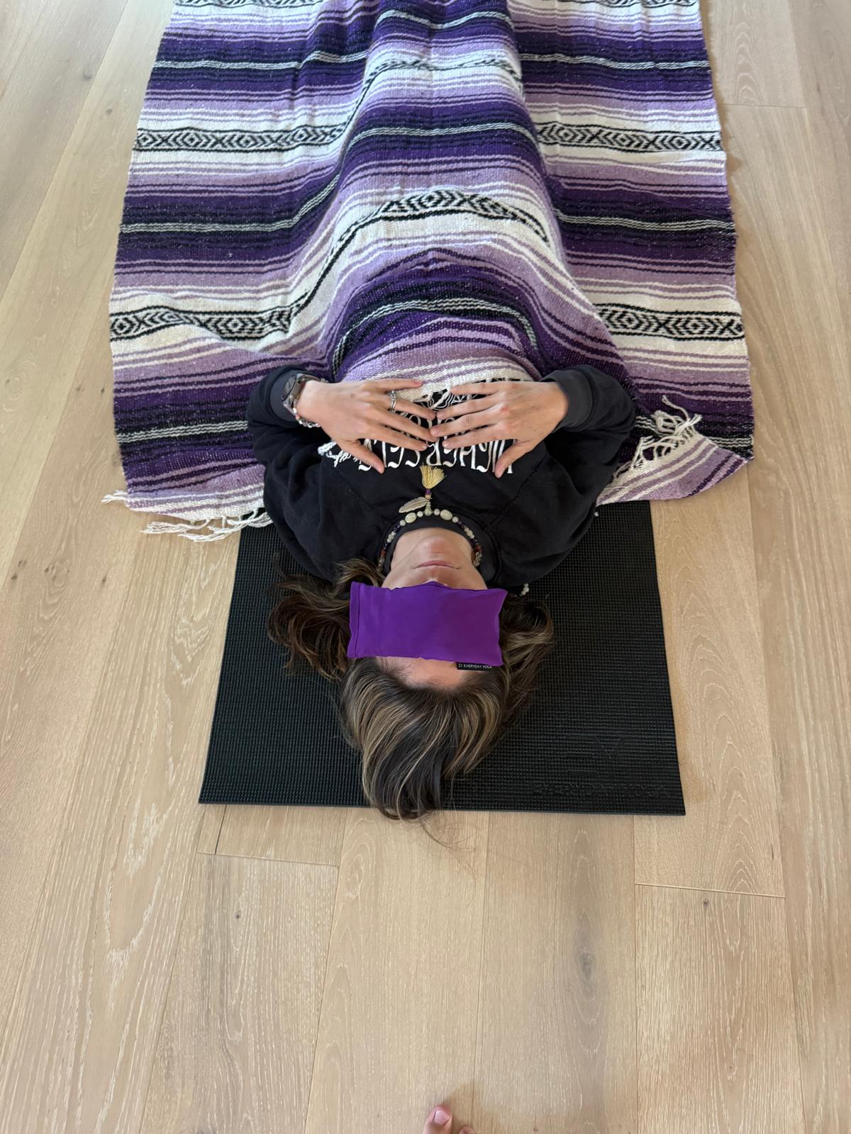 Person lying on a black yoga mat with a purple cloth covering their eyes, on a wooden floor, with a striped blanket above them.