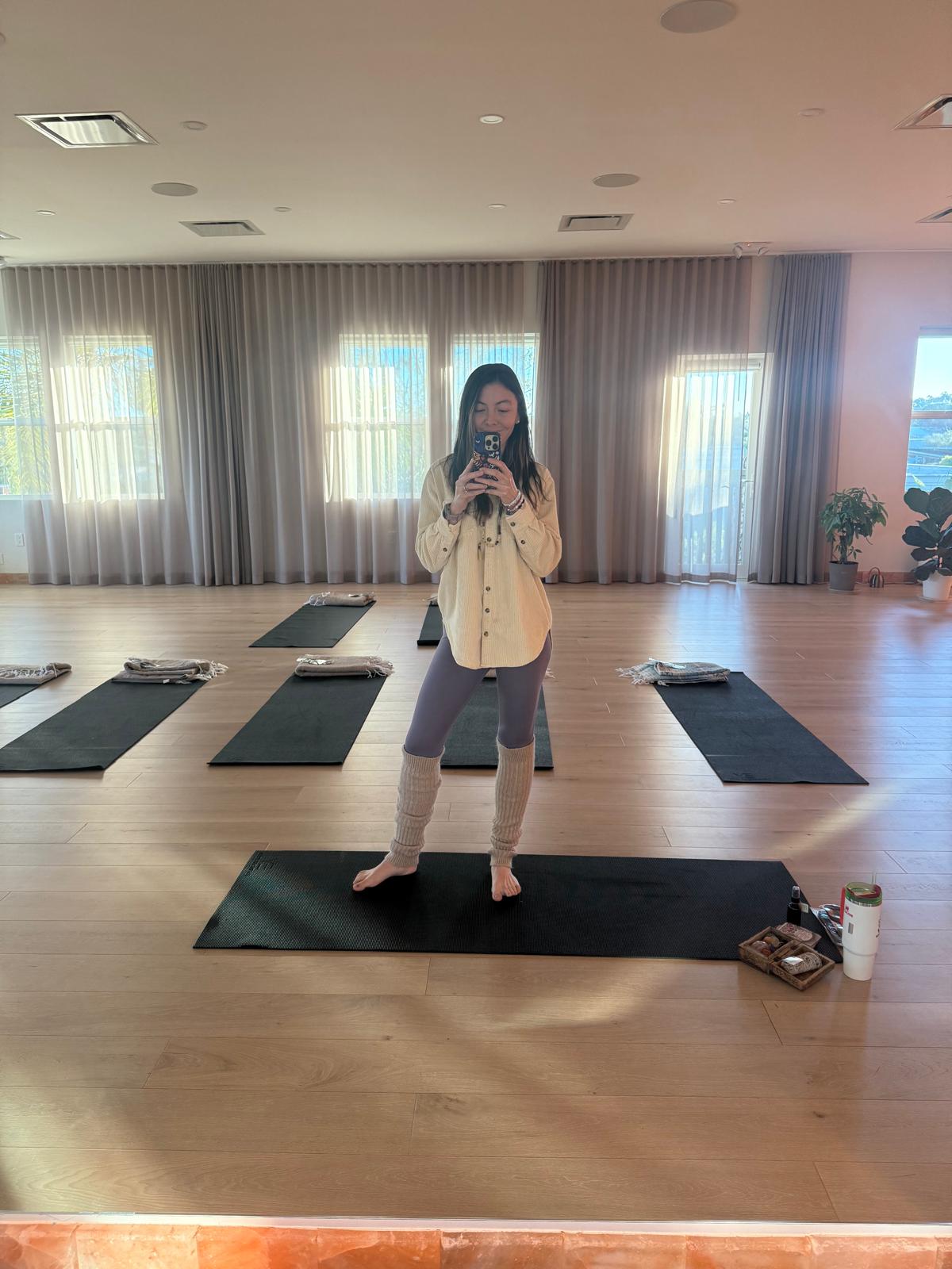 Person taking a mirror selfie in a spacious room with large windows and curtains, standing on a yoga mat.