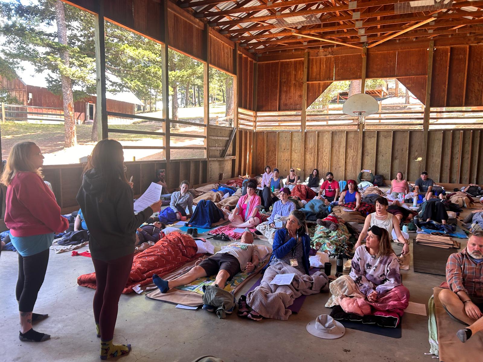 Group of children lying on mats inside a wooden gymnasium, with two women standing and reading aloud, large windows showing trees outside.