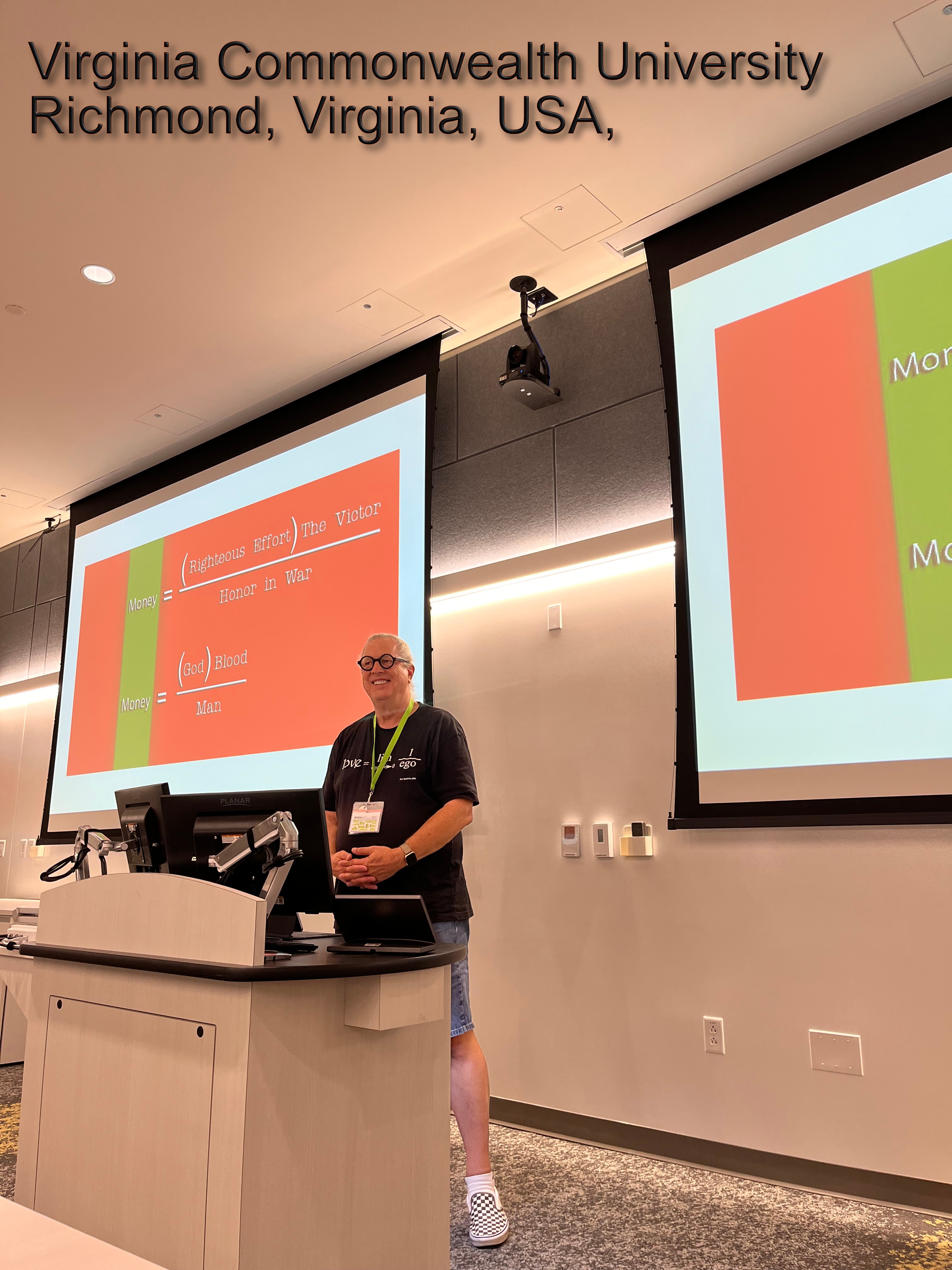 A person stands at a podium in a lecture hall with two large screens displaying a presentation. The room is at Virginia Commonwealth University, Richmond, Virginia, USA.