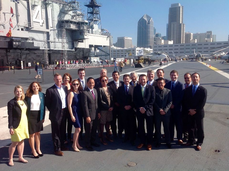 Group of 16 people standing on a ship's deck with a city skyline in the background.