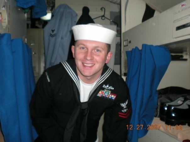 Person in navy uniform smiling, wearing a white sailor hat, in a room with blue curtains and clothing.