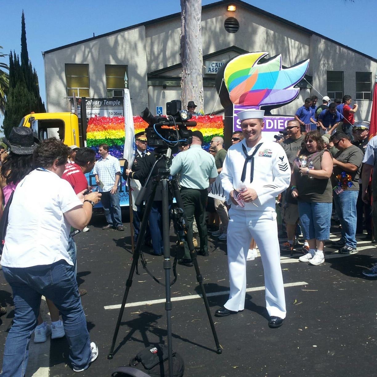 Person dressed in a sailor costume standing in front of a crowd at a pride event with rainbow flags and a colorful boat sculpture.