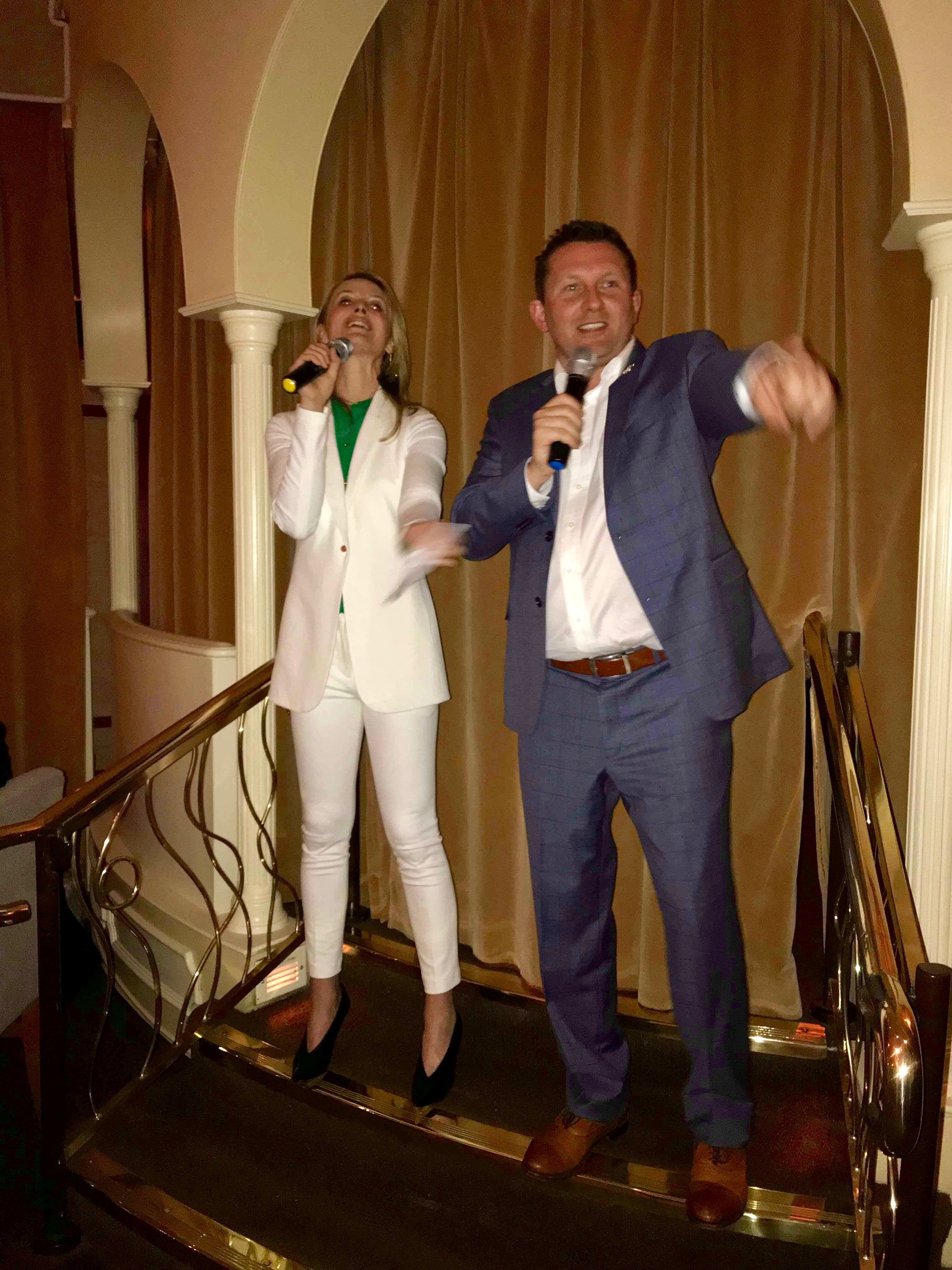 Two people on a staircase, one holding a microphone and smiling, the other drinking from a glass, with a curtain backdrop.