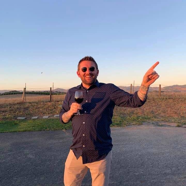 Man with sunglasses holding a glass of wine, smiling outdoors during sunset, pointing to the right, with a fence and landscape in background.