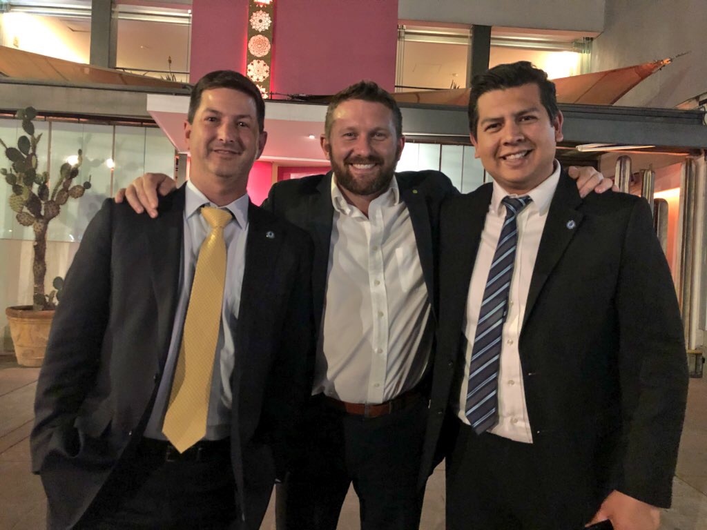Three men in suits smiling with arms around each other indoors, background with plants and pink walls.