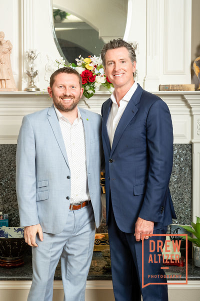 Two men in suits standing in front of a fireplace with a mirror and flowers, smiling at the camera.