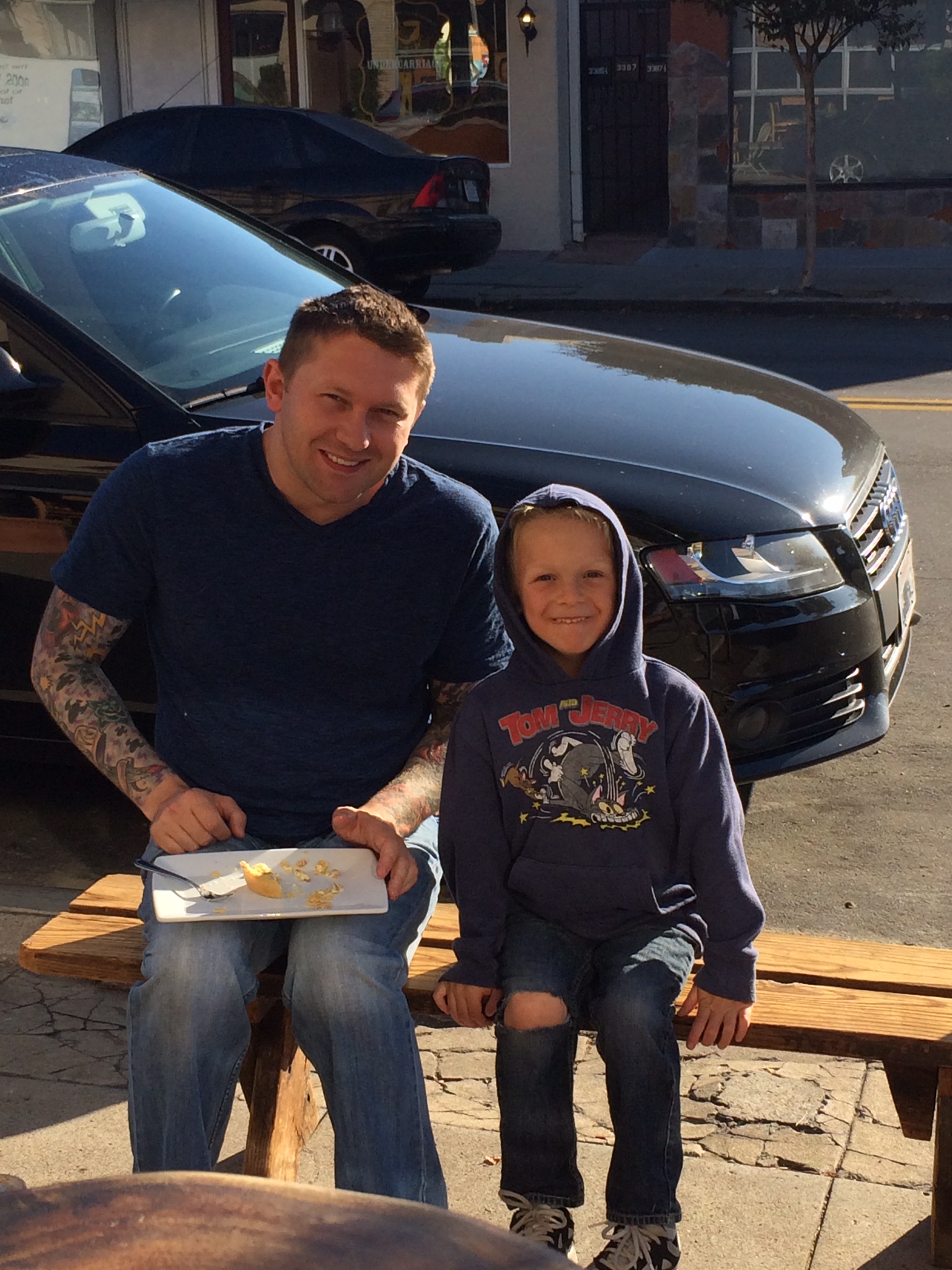 Man and child sitting on a bench outdoors, smiling, with a black car behind them, sunny day.