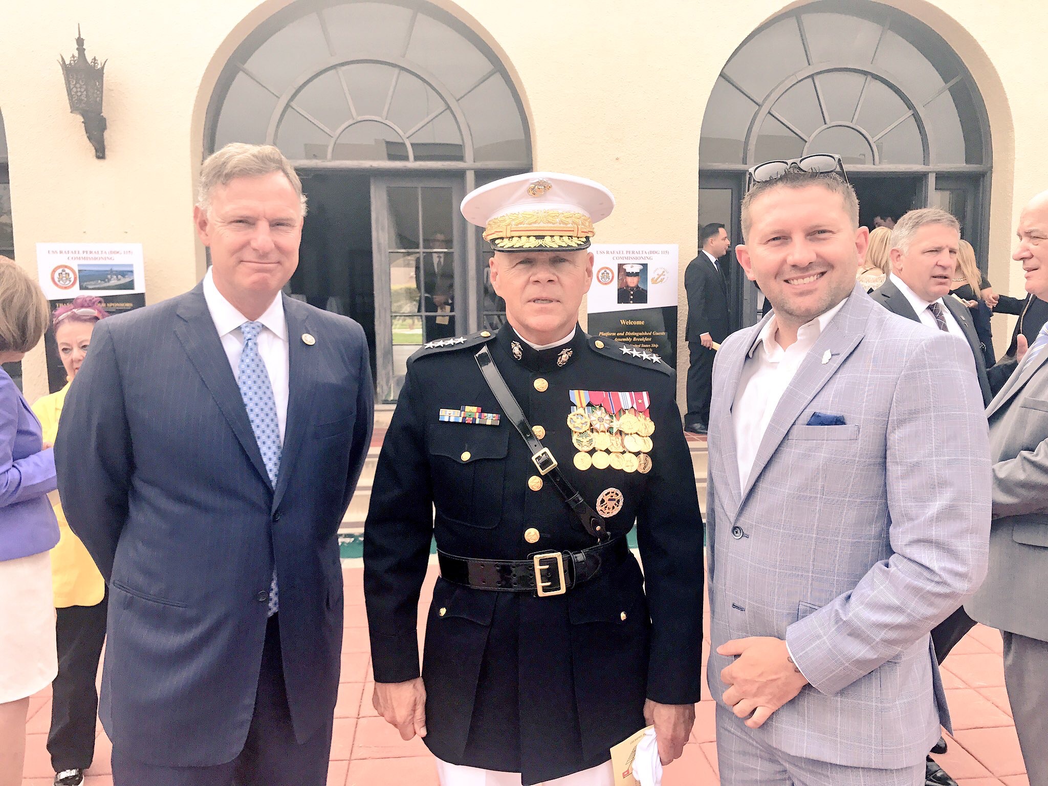 Three men in formal attire standing outside a building with arched windows, one in military uniform, two in suits.