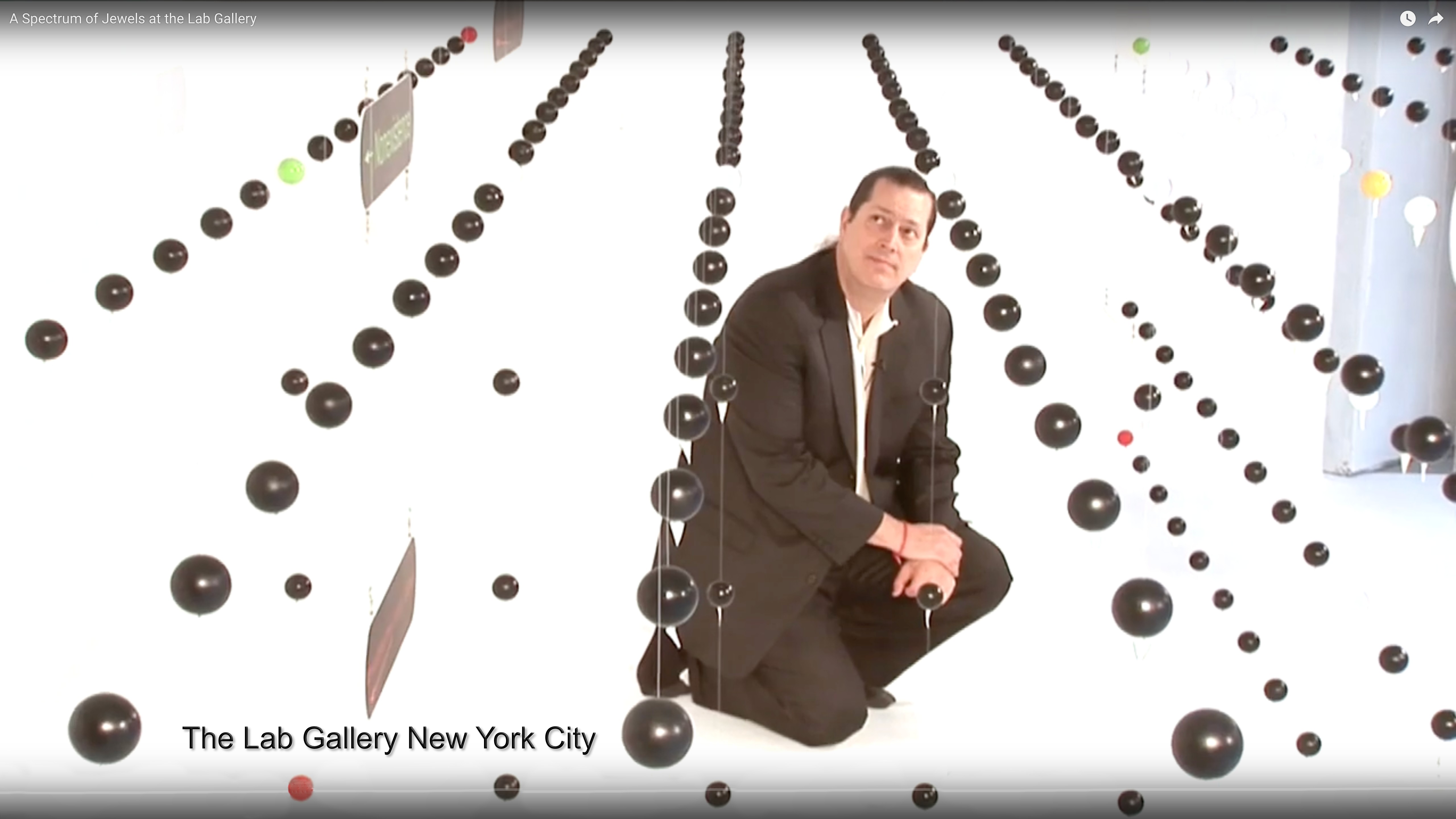 Man in black suit crouching among black and white spheres in an art gallery, with text 'The Lab Gallery New York City' at bottom.