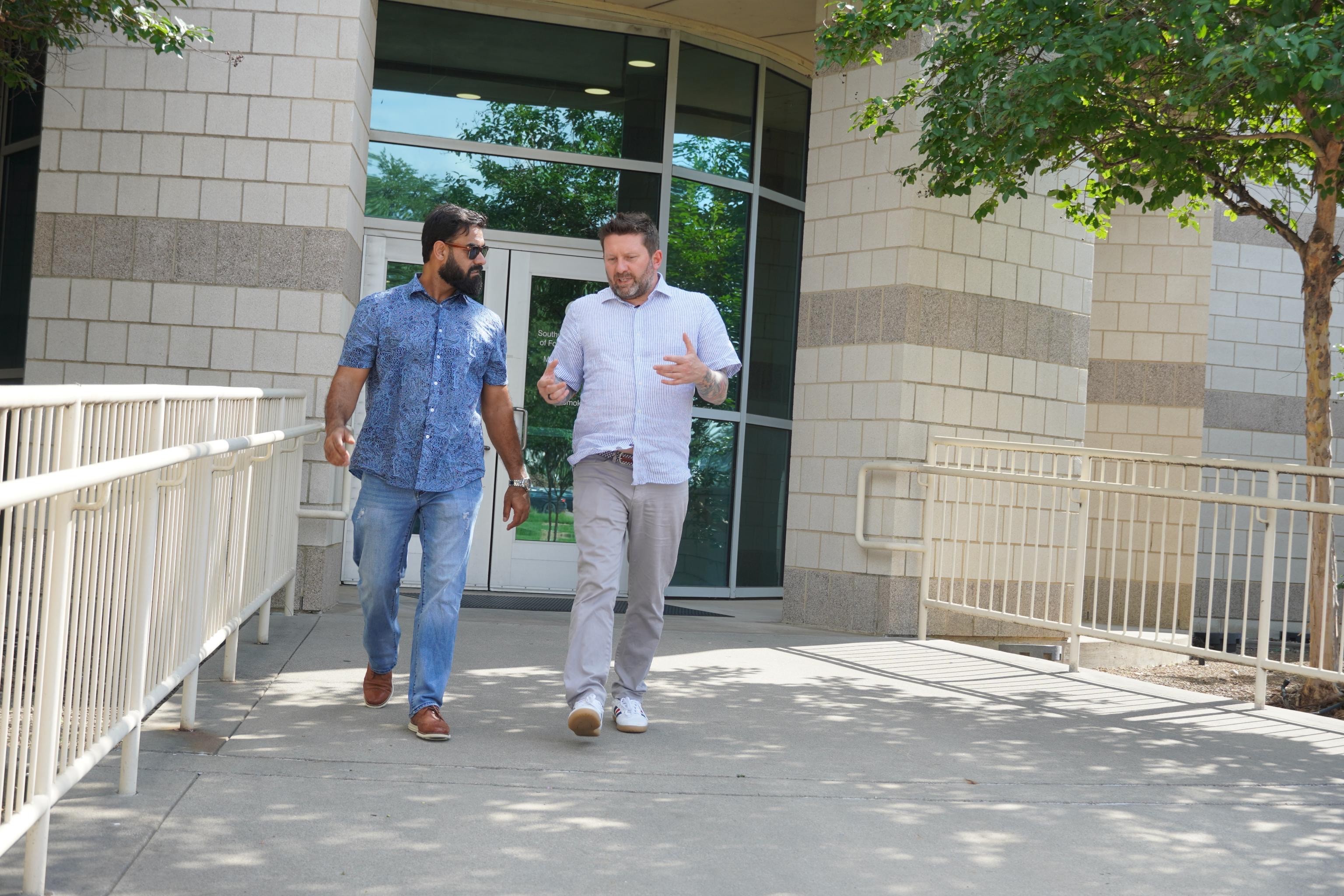 Two men walk and talk outside a modern building with glass doors, surrounded by trees and railings.