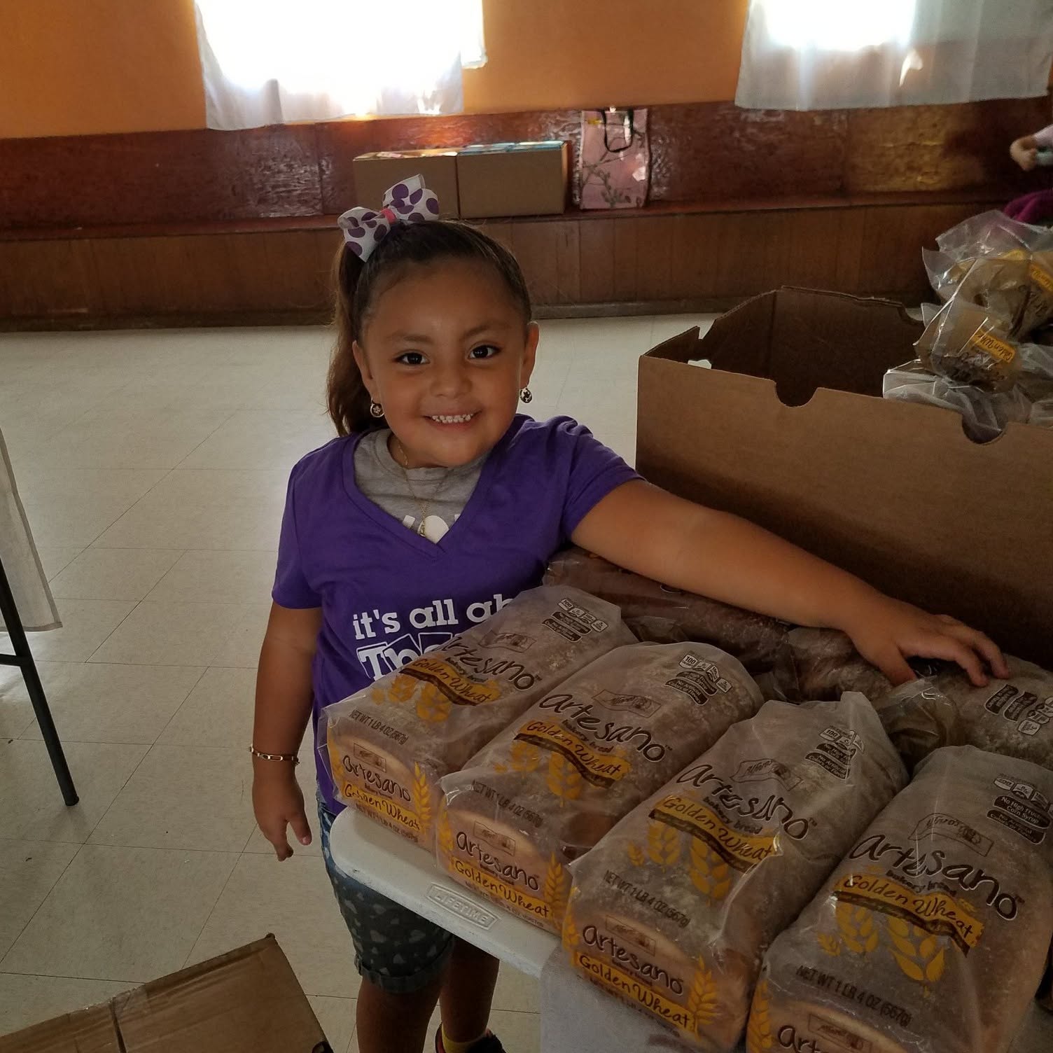 Smiling young girl with a purple shirt and a bow in her hair, reaching for bread on a table.