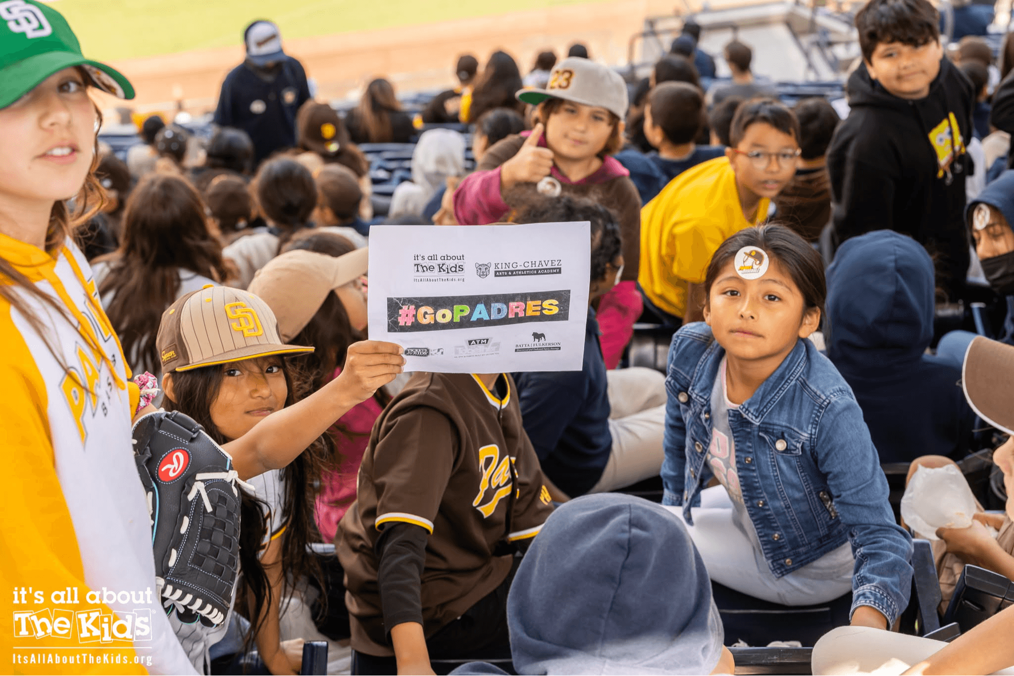 Group of children and adults outdoors, some holding a sign with hashtag #GoPADRES, sitting and standing, wearing casual clothing and hats.