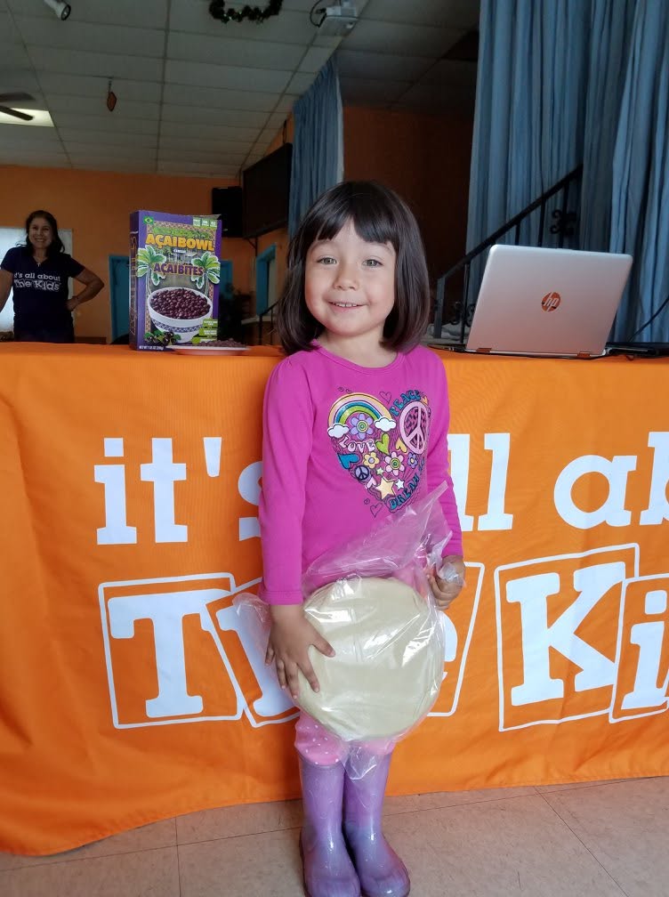 Young girl with dark hair and a pink shirt holding a large round object, standing in front of an orange table with a TV logo.