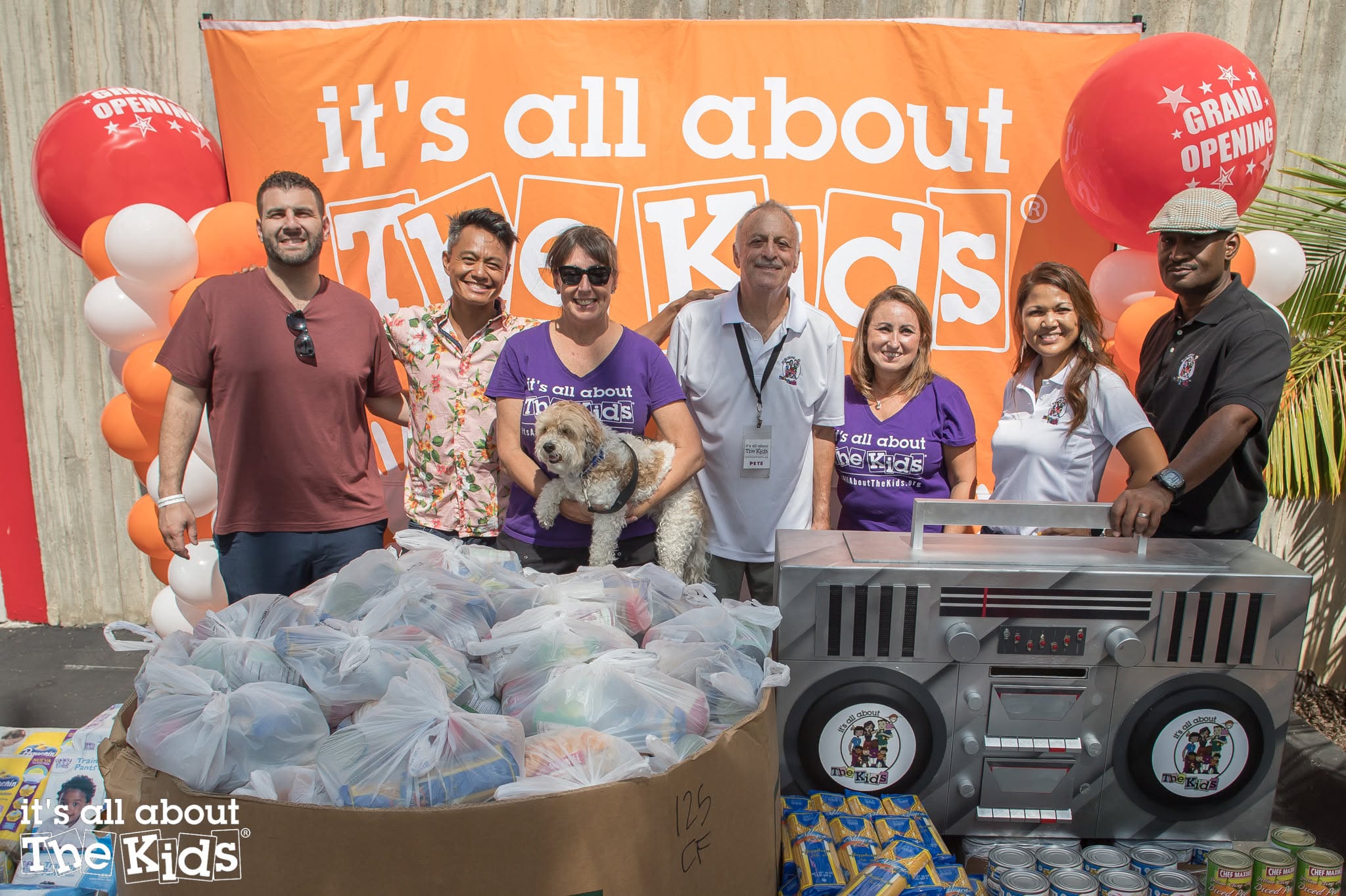 Group of seven people standing outdoors behind a large box of bags, with balloons and a banner reading 'it's all about Kids'.