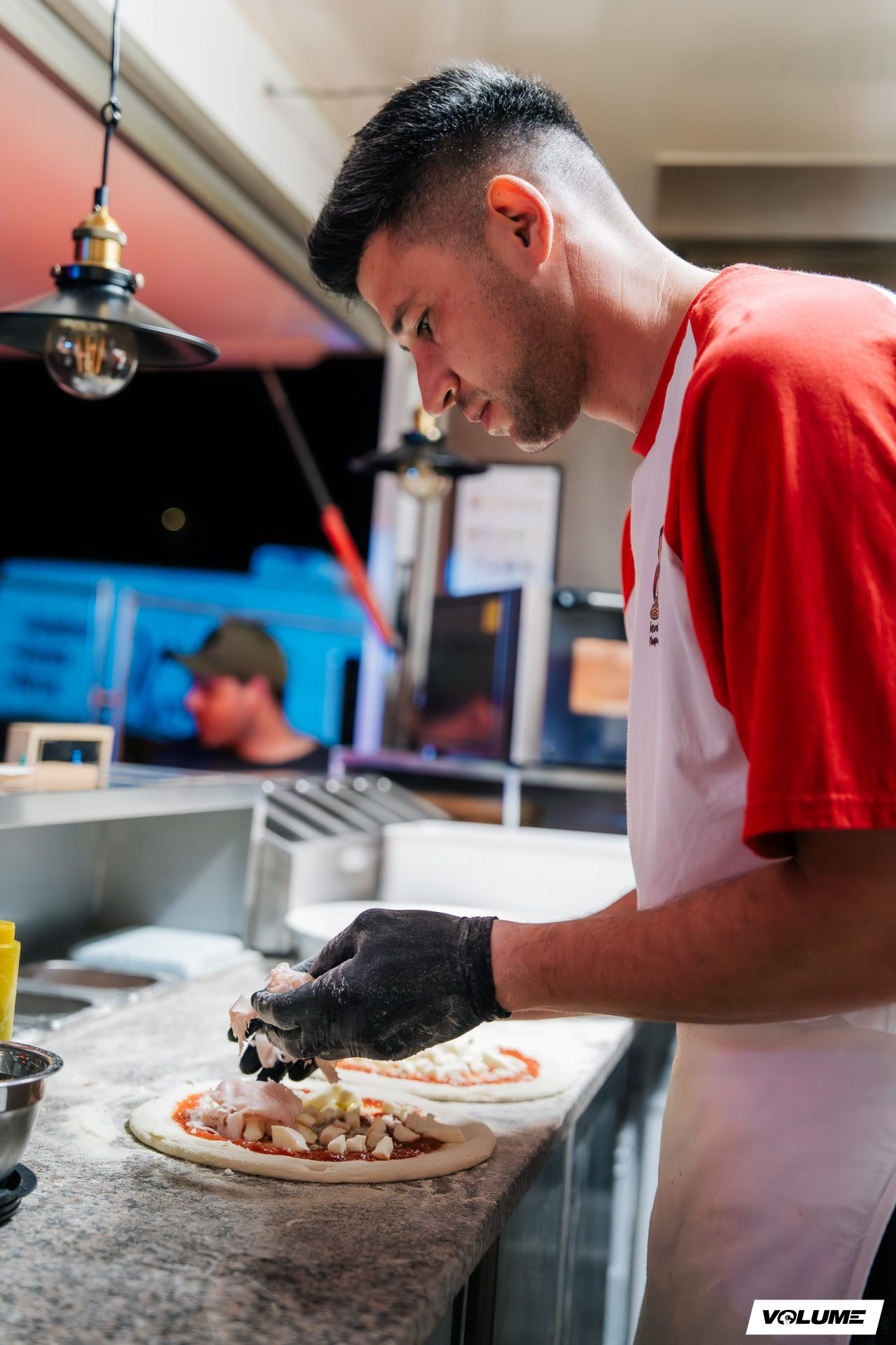 Young man preparing pizza in a kitchen, wearing a red and white shirt, with a focused expression, and another person in the background.