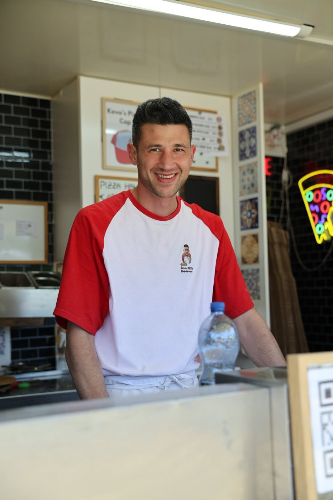 Smiling man in a red and white shirt behind a counter with a water bottle, in a fast-food setting.