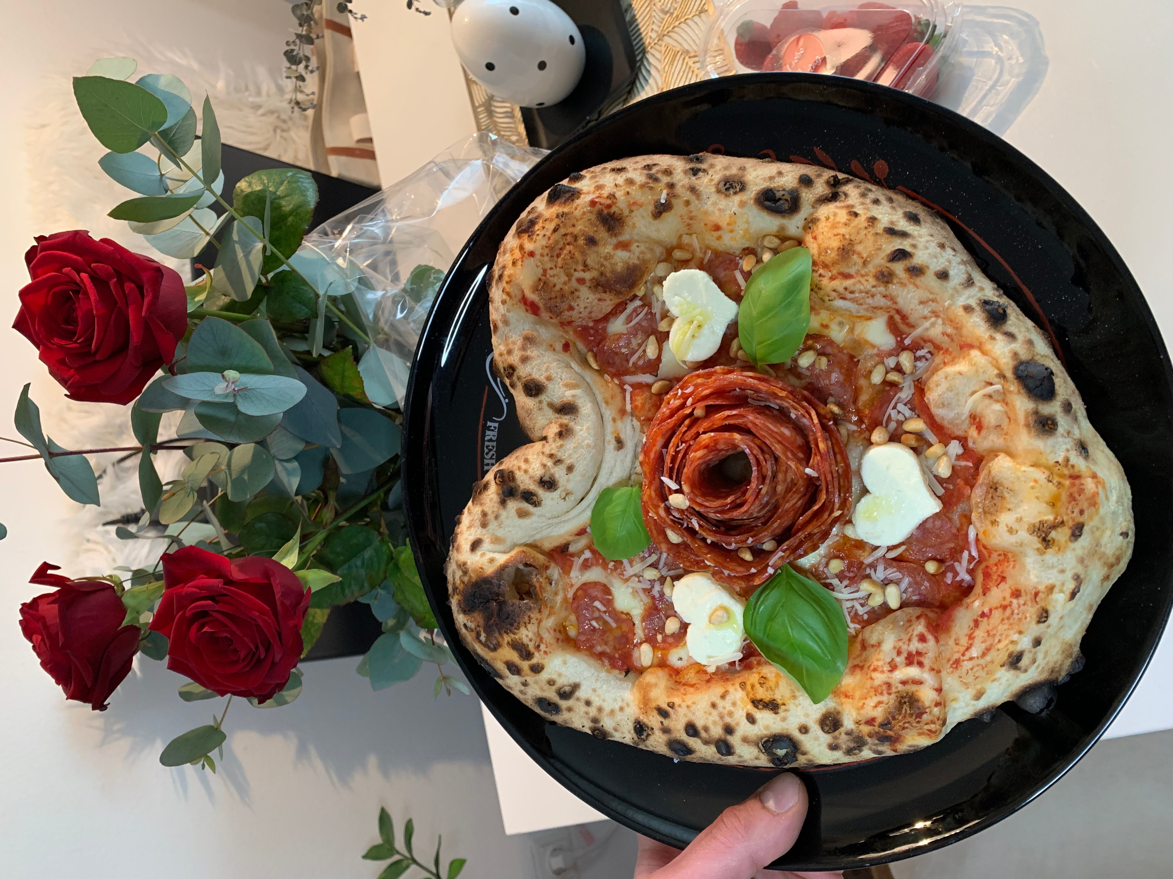 Heart-shaped pizza with basil, cheese, and pepperoni, garnished with a rose-shaped topping, on a black plate, next to red roses.