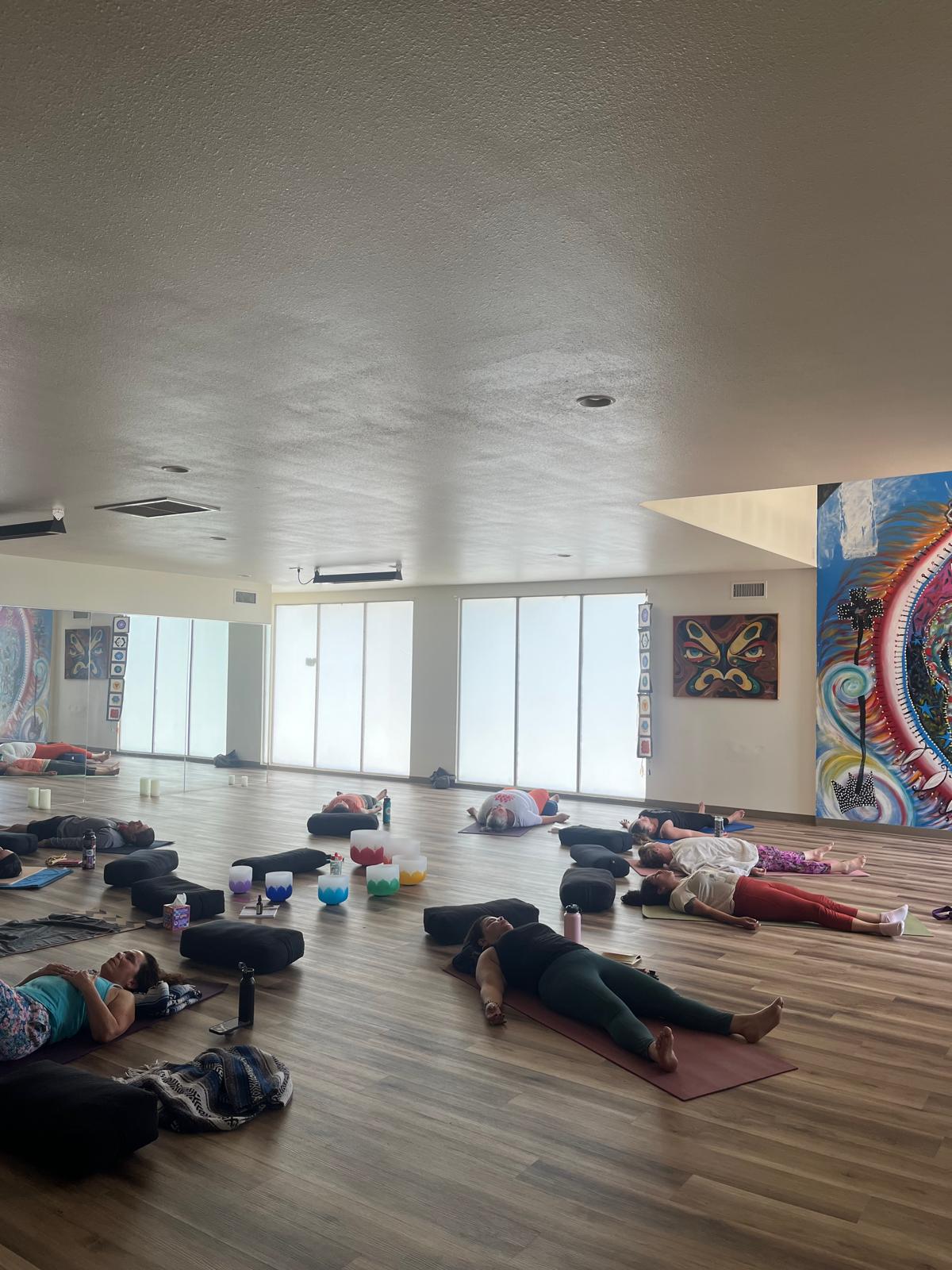 People lying on mats in a spacious room with large windows and colorful artwork on the walls.