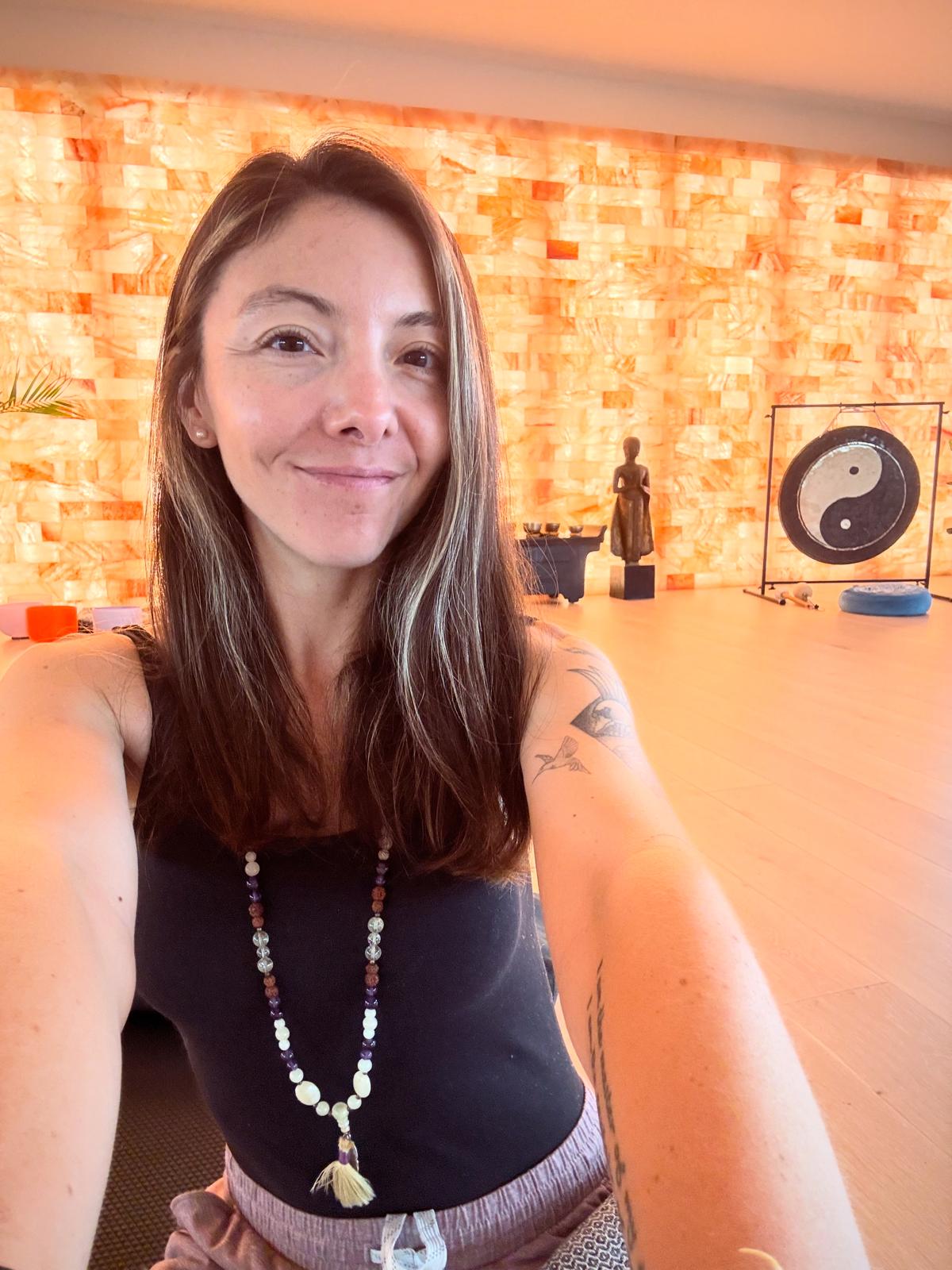 Woman with long hair smiling, wearing a black top and beaded necklace, in a room with brick wall and yin-yang symbol.