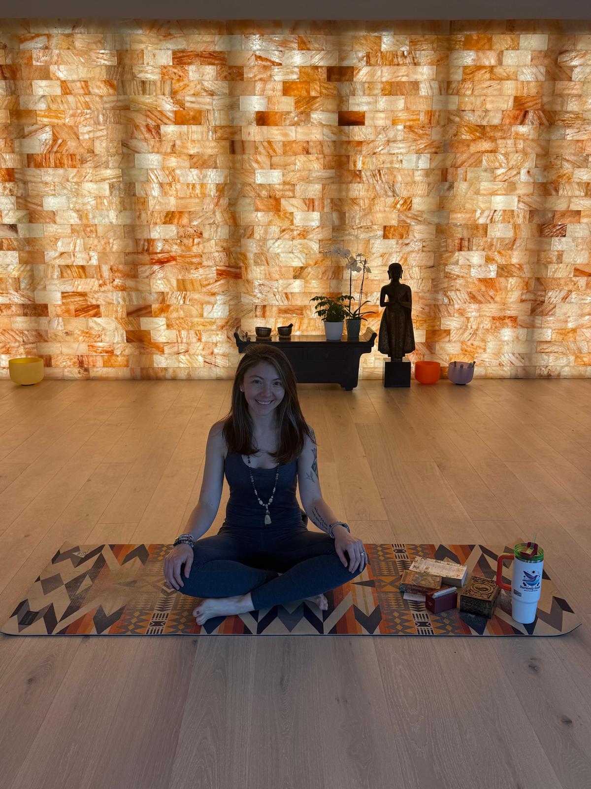 Person sitting cross-legged on a patterned mat in front of a brick wall with decorative objects and a statue behind.