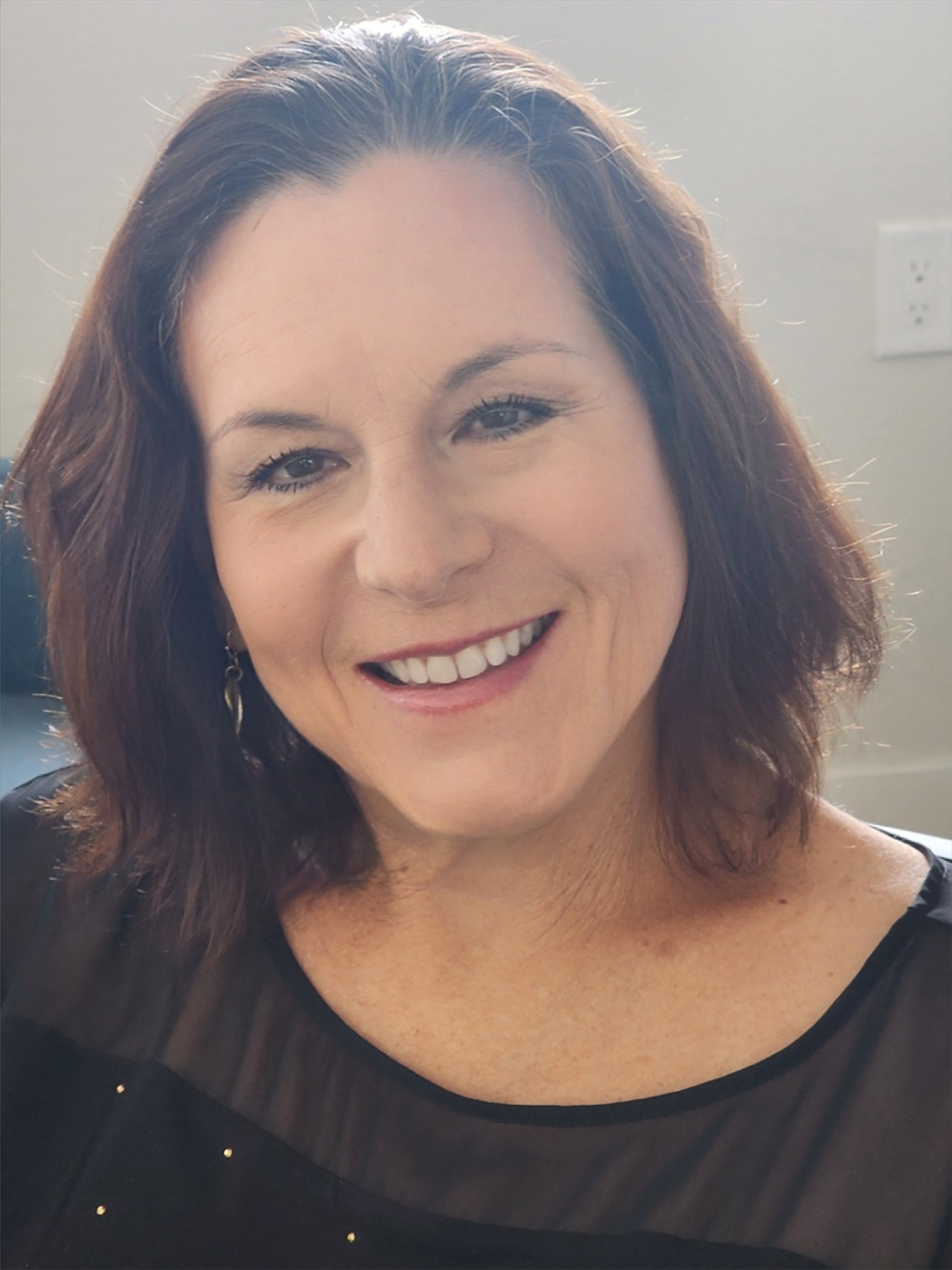 Smiling woman with shoulder-length dark hair, wearing a black top, indoors near a wall outlet.