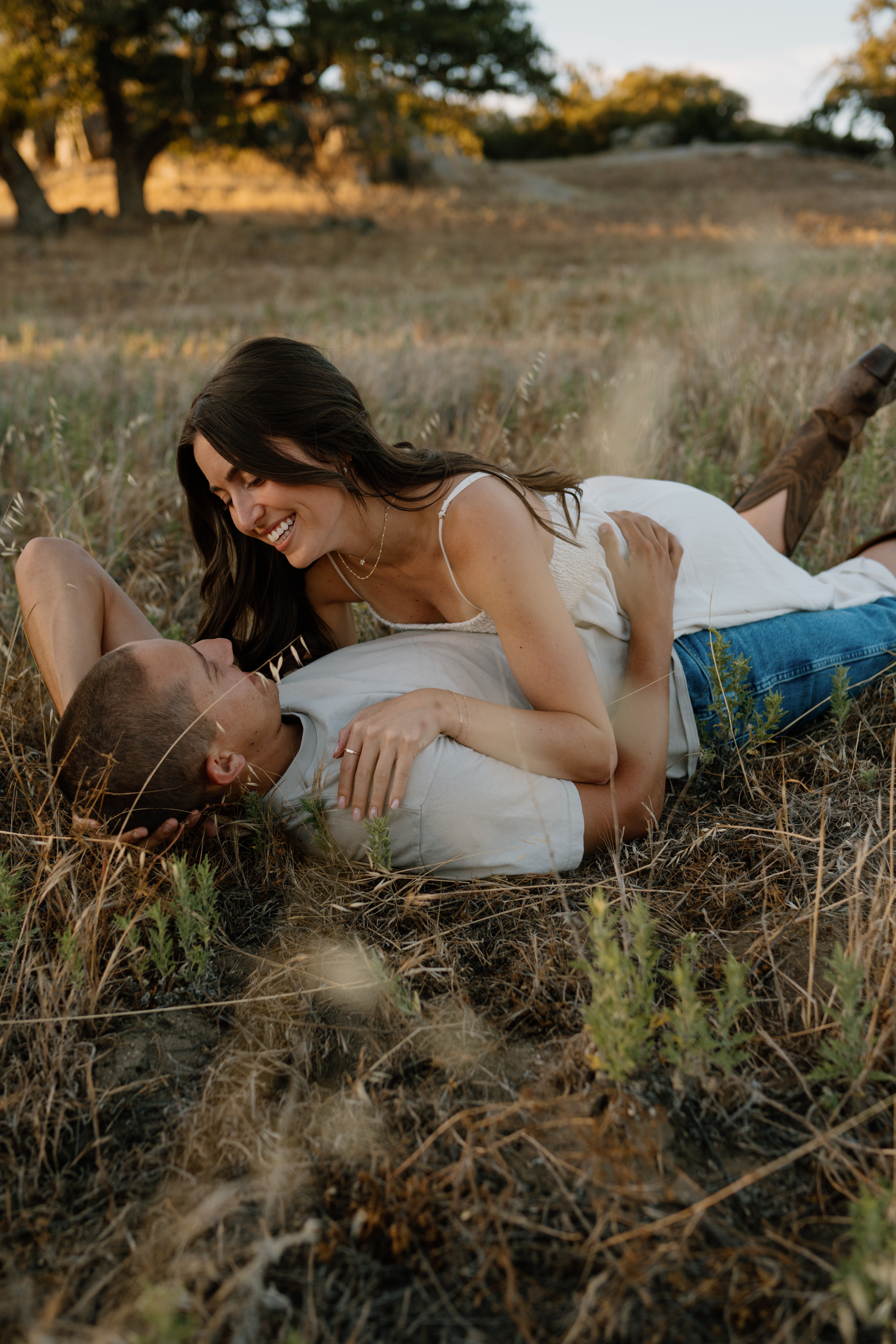 A woman and a man lying on the grass outdoors, smiling and looking at each other, with trees and open land in the background.