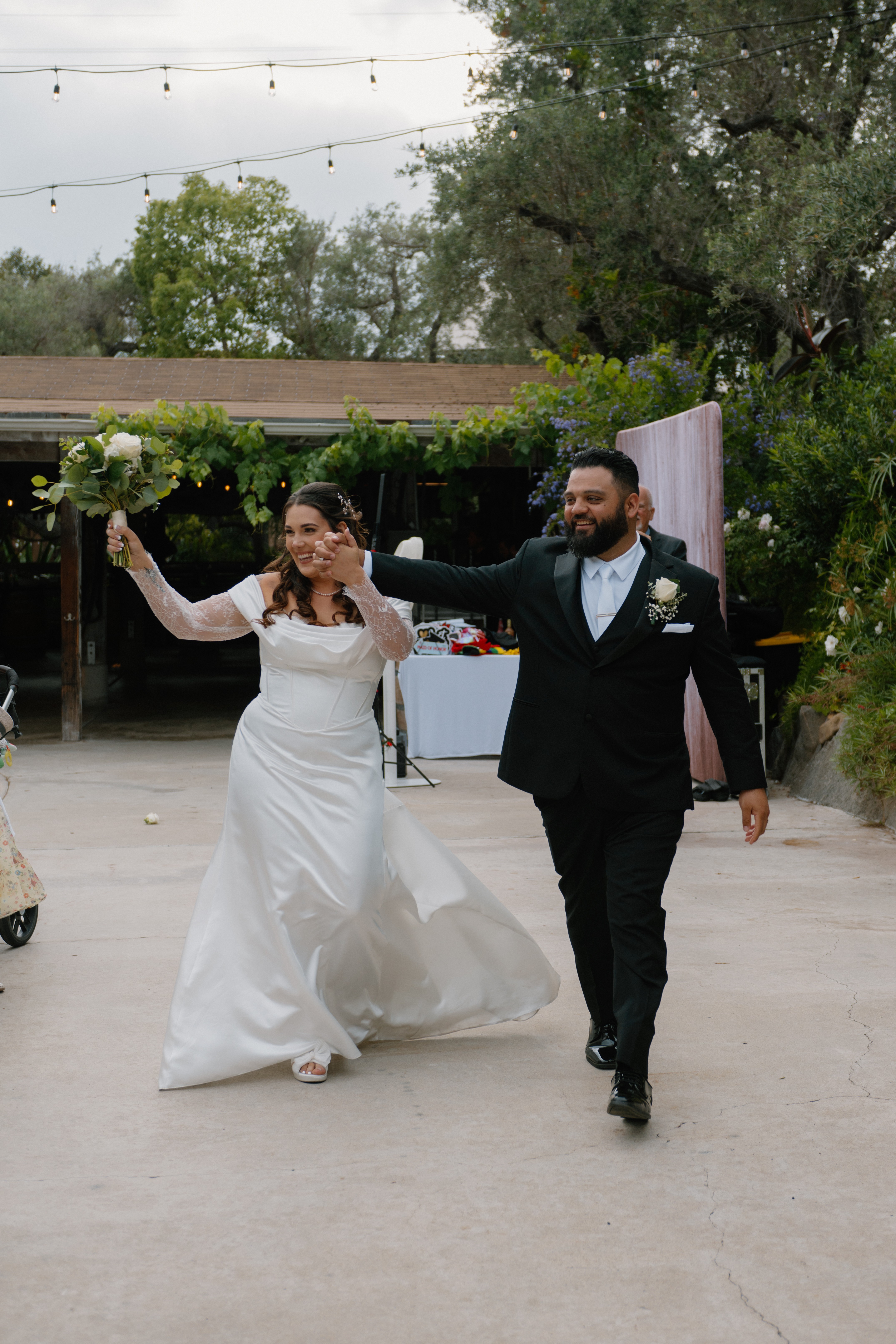 Bride and groom walking outdoors, holding hands, smiling, bride in white dress, groom in black suit, trees and sky in background