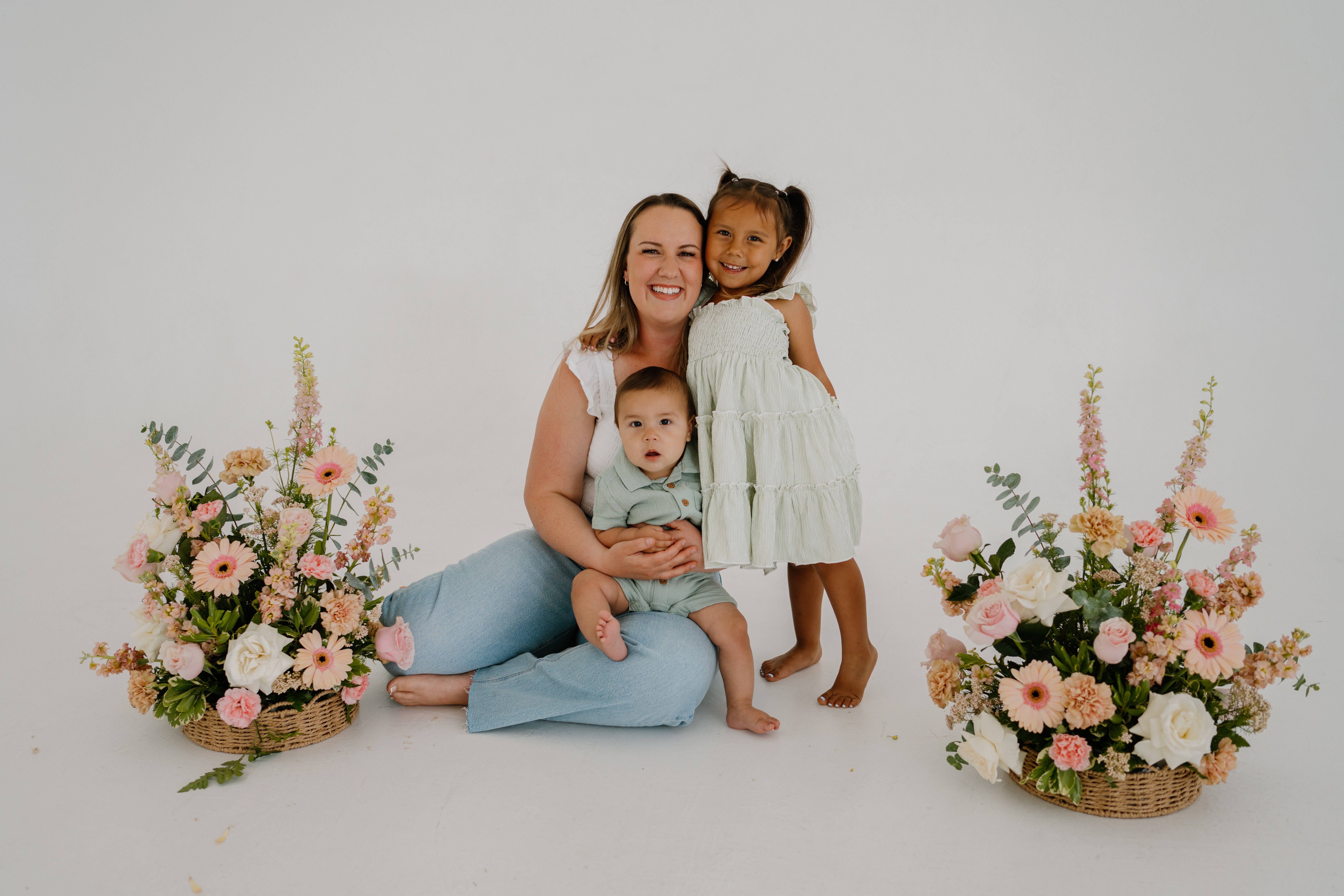 Woman sitting with two children, surrounded by two large floral arrangements, all smiling, against a plain white background.