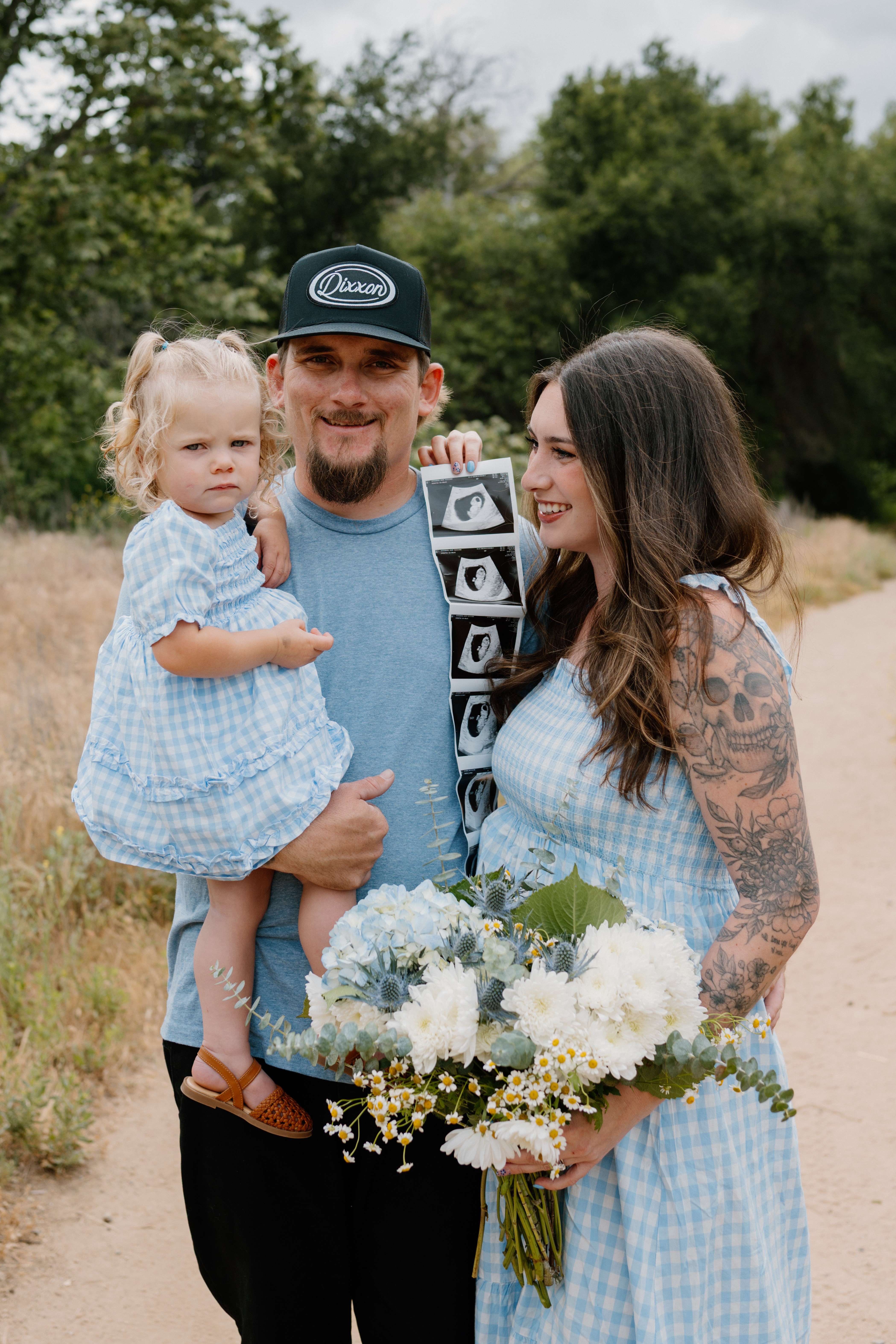 Family of three outdoors, man holding a young girl, woman with a tattooed arm holding flowers, trees in background.