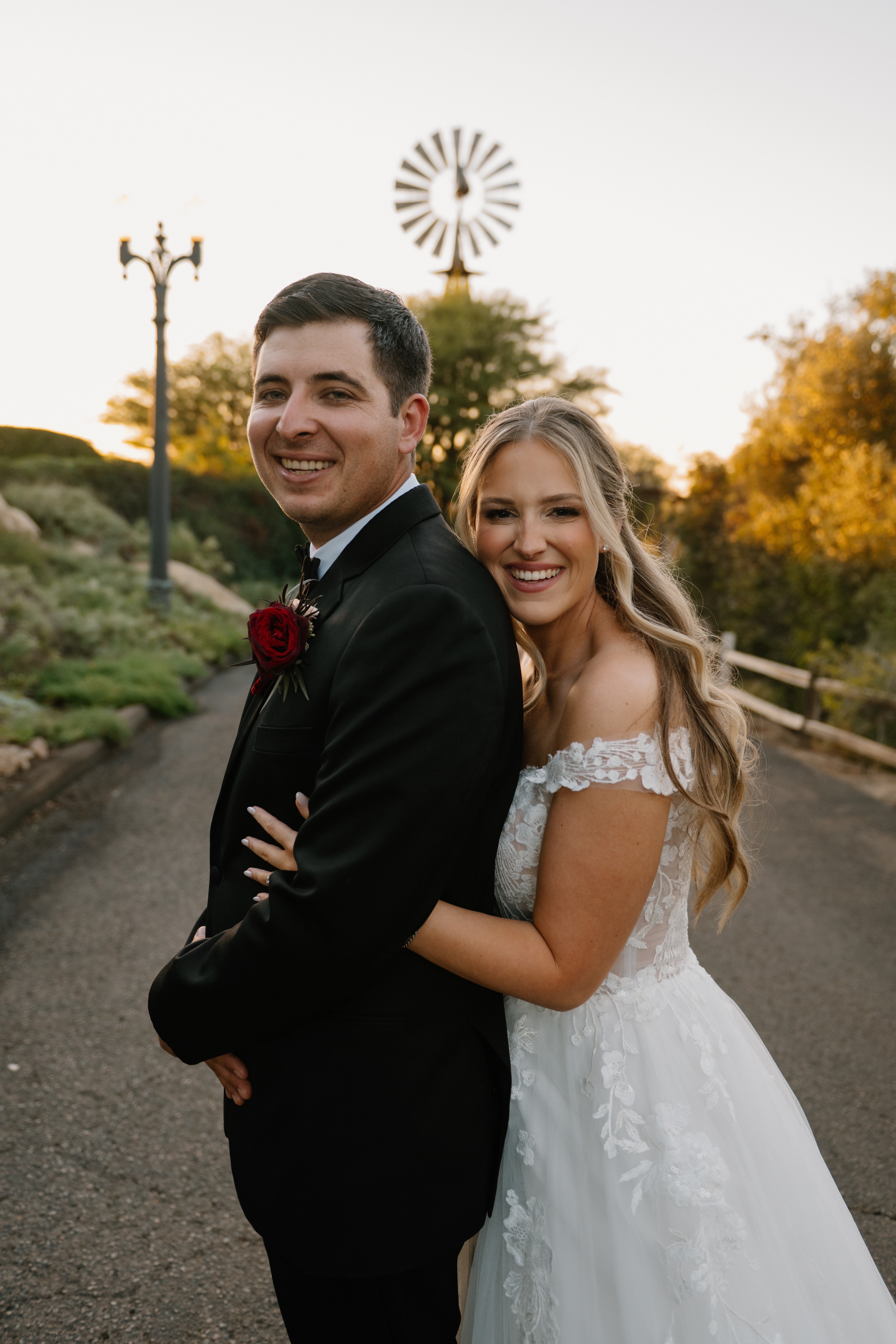 A smiling couple in wedding attire outdoors, with trees and a windmill in the background.