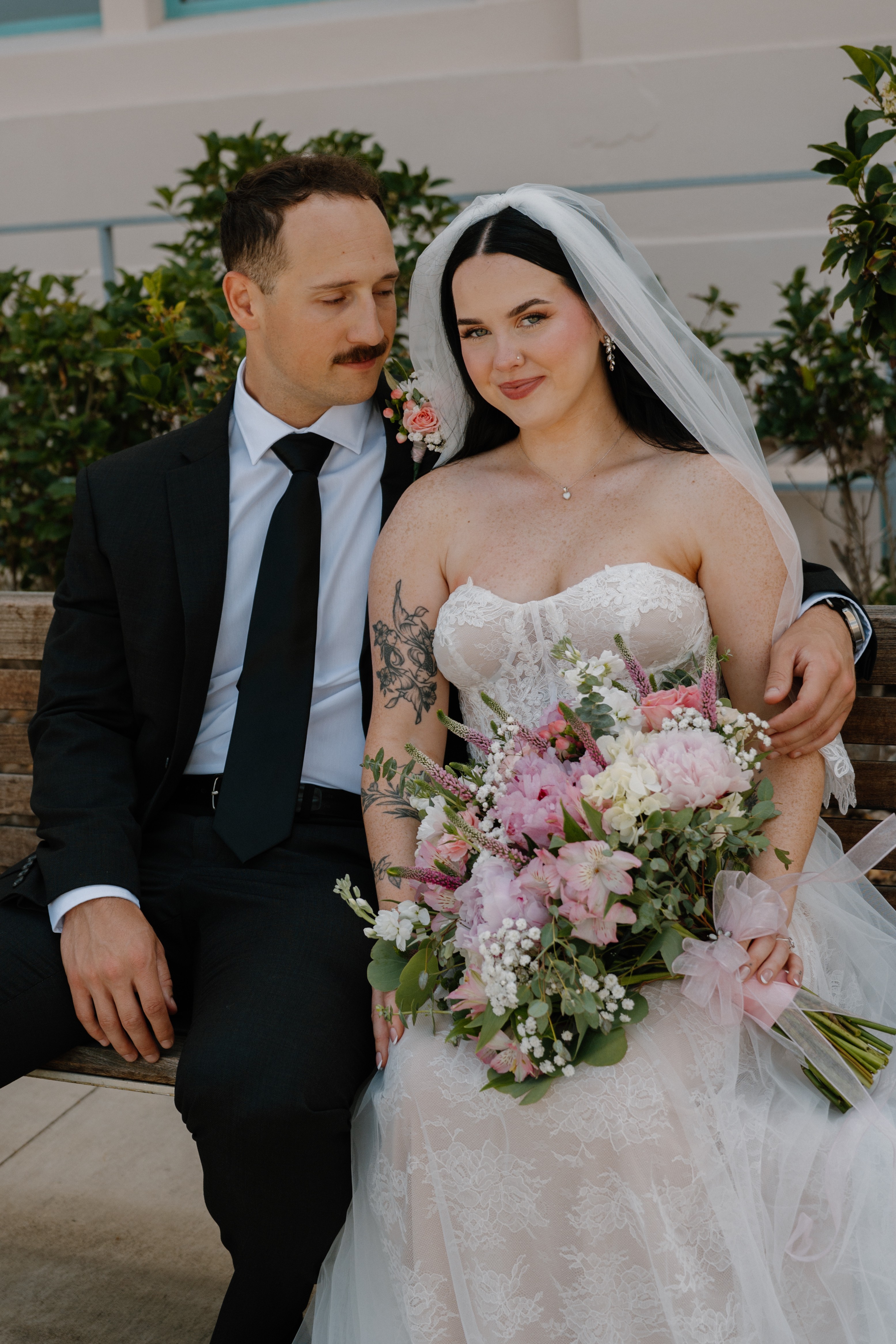 Bride in wedding dress holding a bouquet, seated next to groom in suit, outdoors with greenery in background.