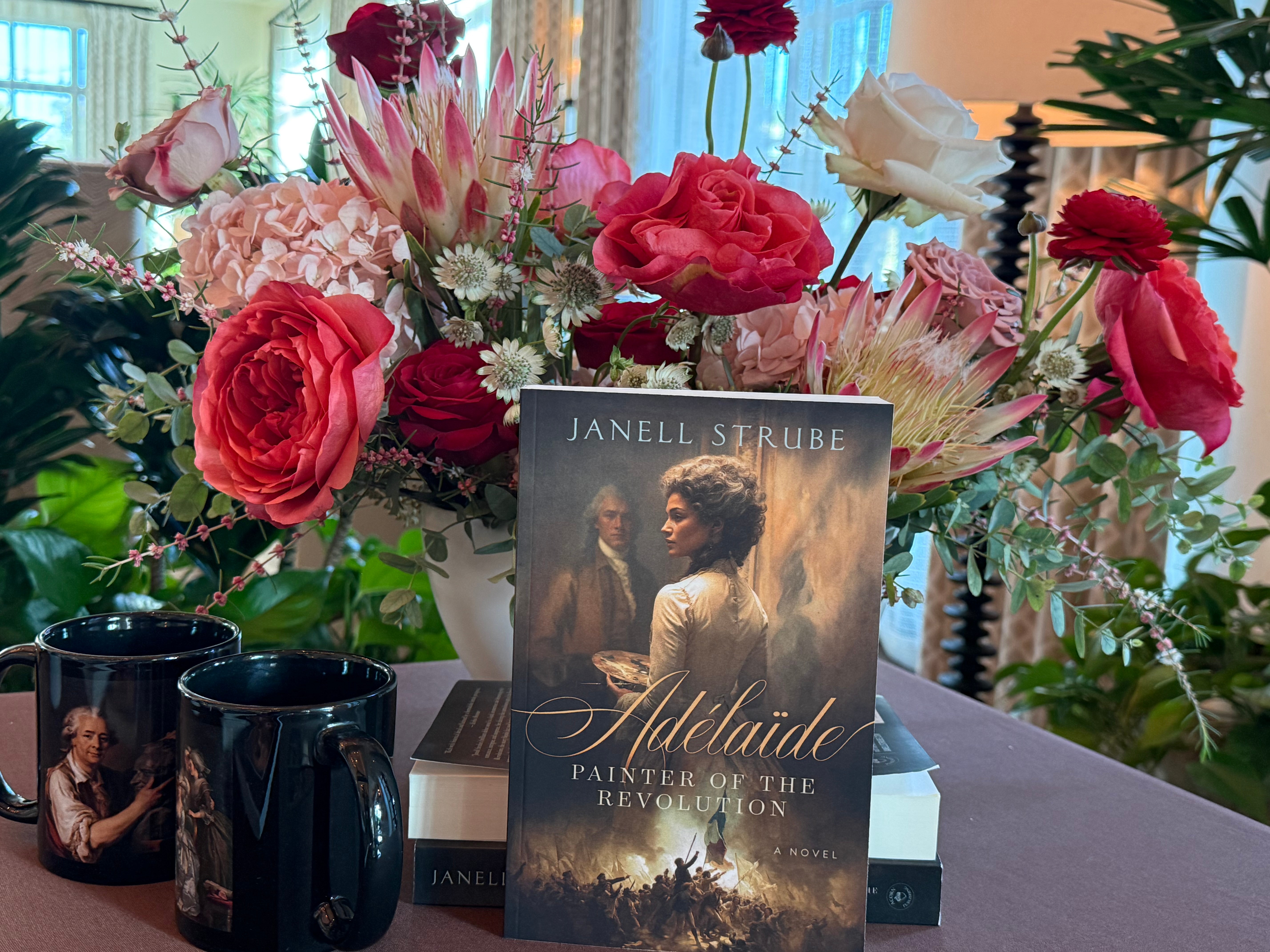 Book titled 'Addled: Painter of the Revolution' placed on a table with a large bouquet of pink, red, and white flowers in the background.