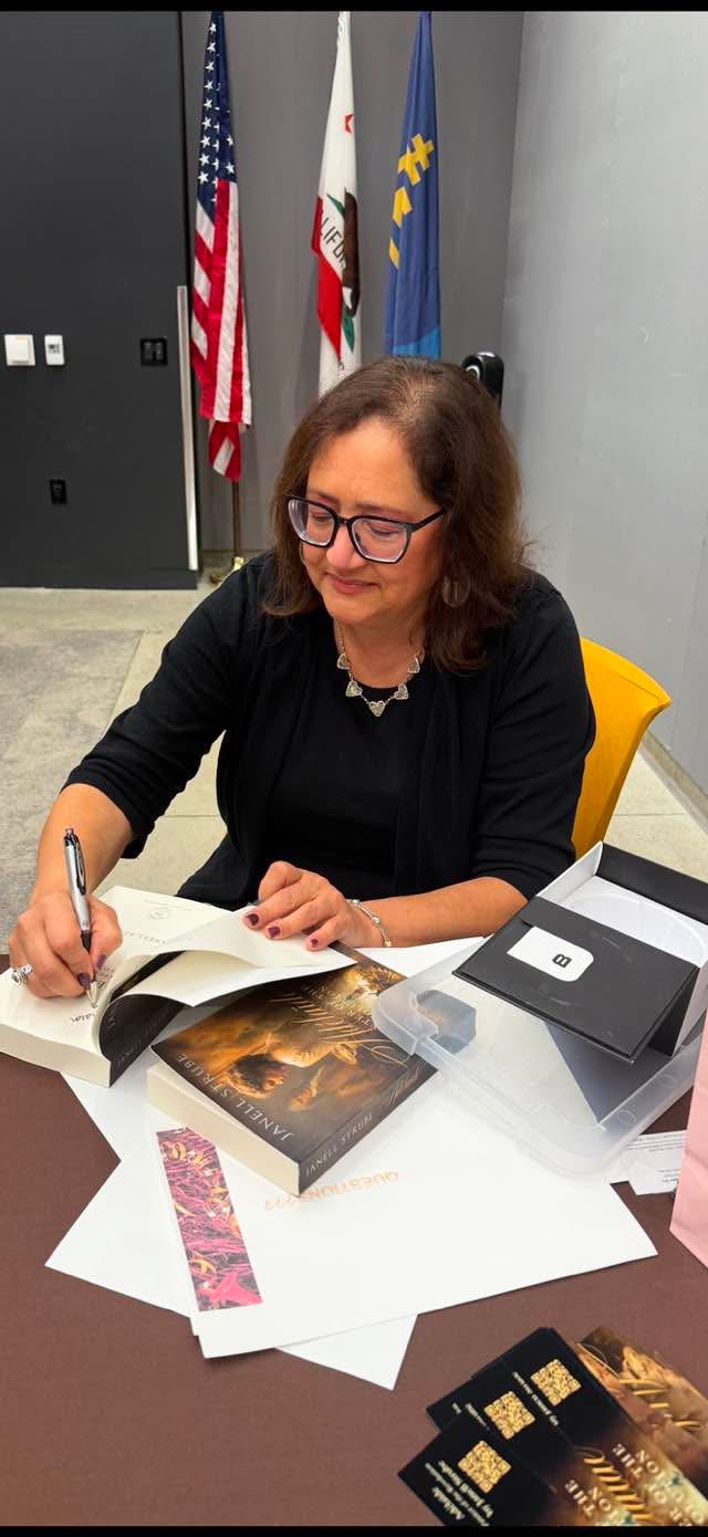 Woman with glasses signing a book at a table with flags in the background.