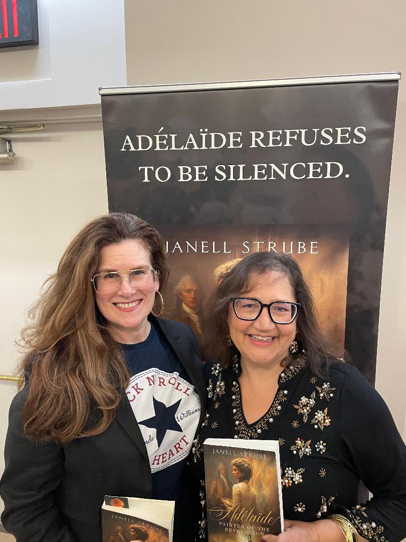 Two women smiling in front of a poster for a book titled 'Adélaïde Refuses to Be Silenced.'