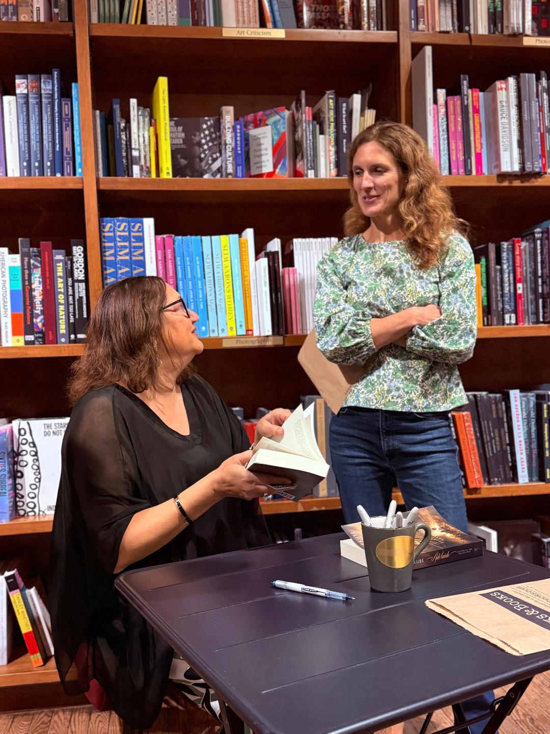 Two women in a library, one sitting at a table with a book, the other standing and smiling, bookshelves in background.